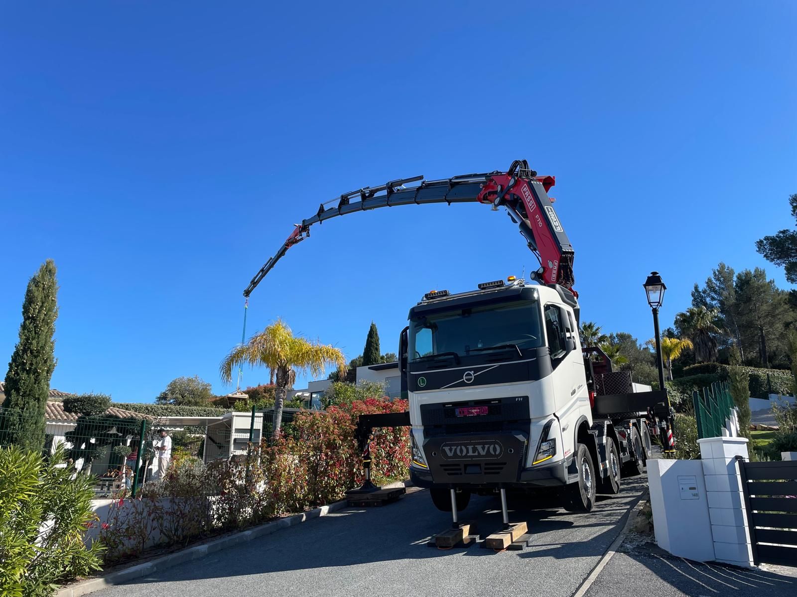 Un camion-grue garé dans une rue ensoleillée, la flèche déployée au-dessus d’une maison et d’un jardin.