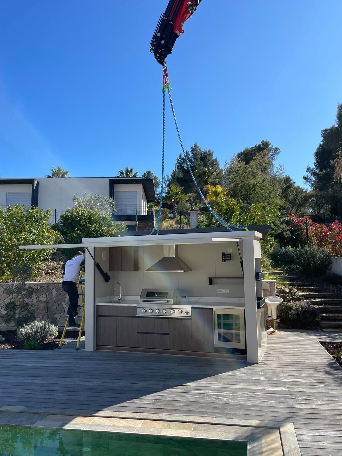 Un îlot de cuisine d’extérieur soulevé par une grue dans un jardin ensoleillé, avec une maison moderne et des arbres.