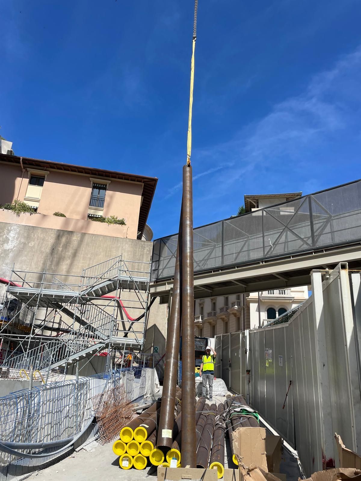 Chantier avec une grue qui soulève un long poteau près d’un bâtiment endommagé et d’un ouvrier portant un gilet jaune