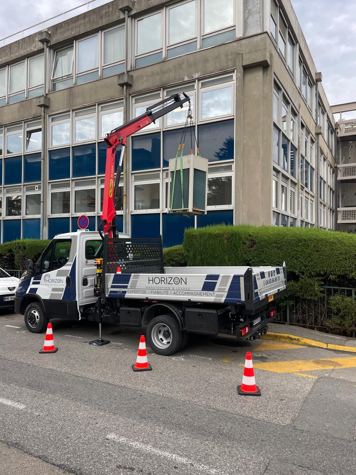 Camion-grue rouge garé près d’un bâtiment, avec des cônes de signalisation et une haie le long de la rue