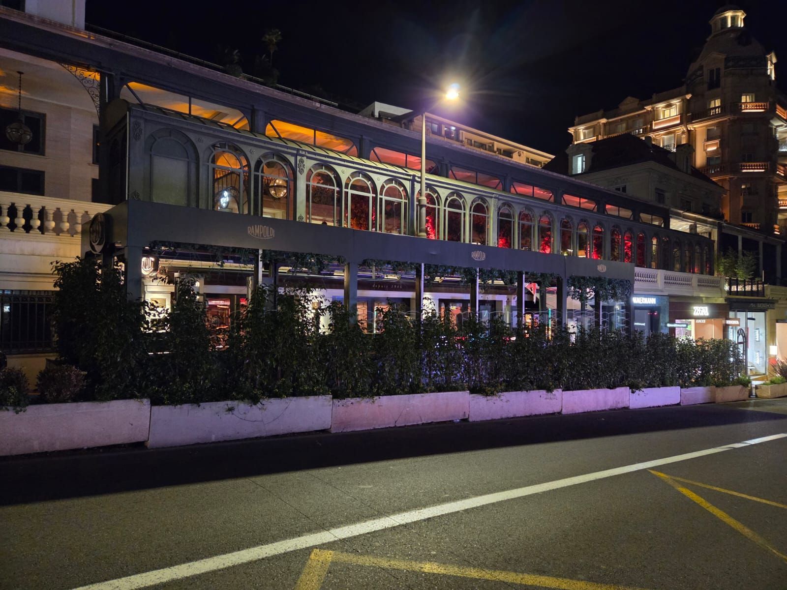 Vue nocturne d’une rue avec un restaurant éclairé, des auvents rouges et une tour sur le toit en arrière-plan