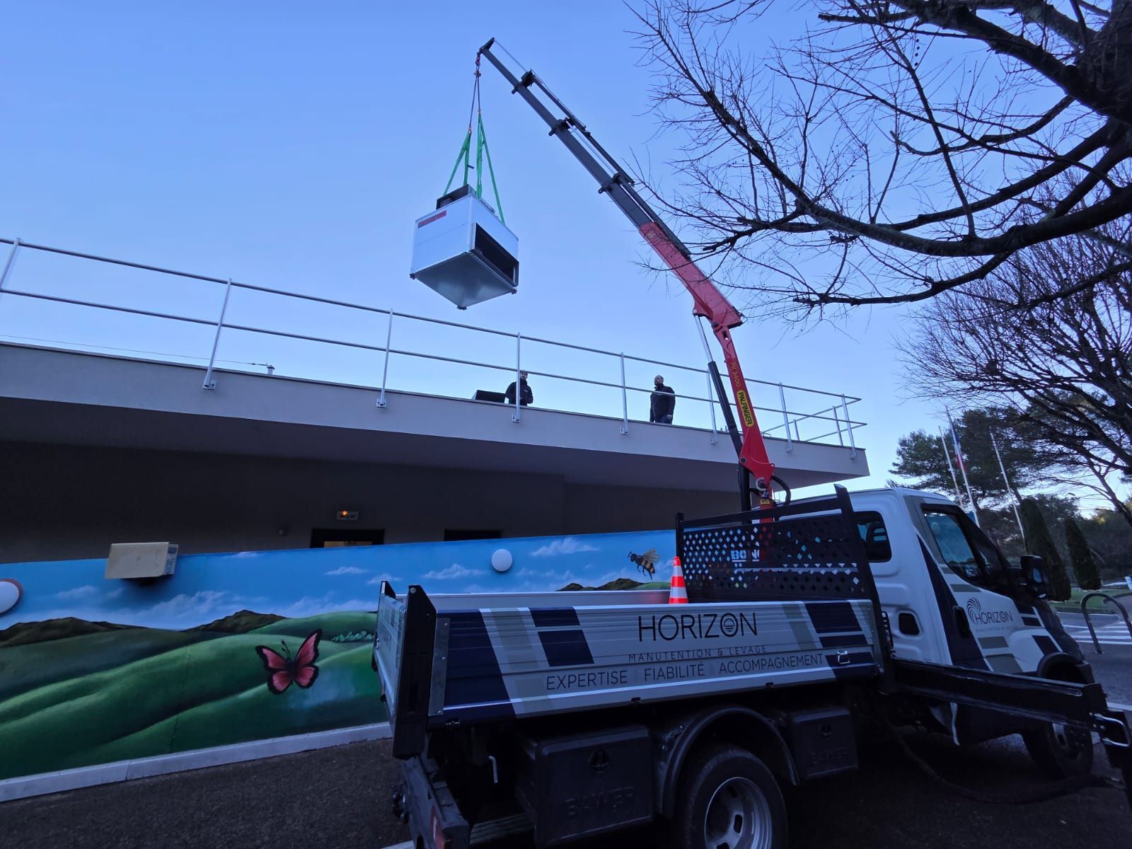 Une grue mobile soulève un cube suspendu au-dessus d’un viaduc à la tombée du jour