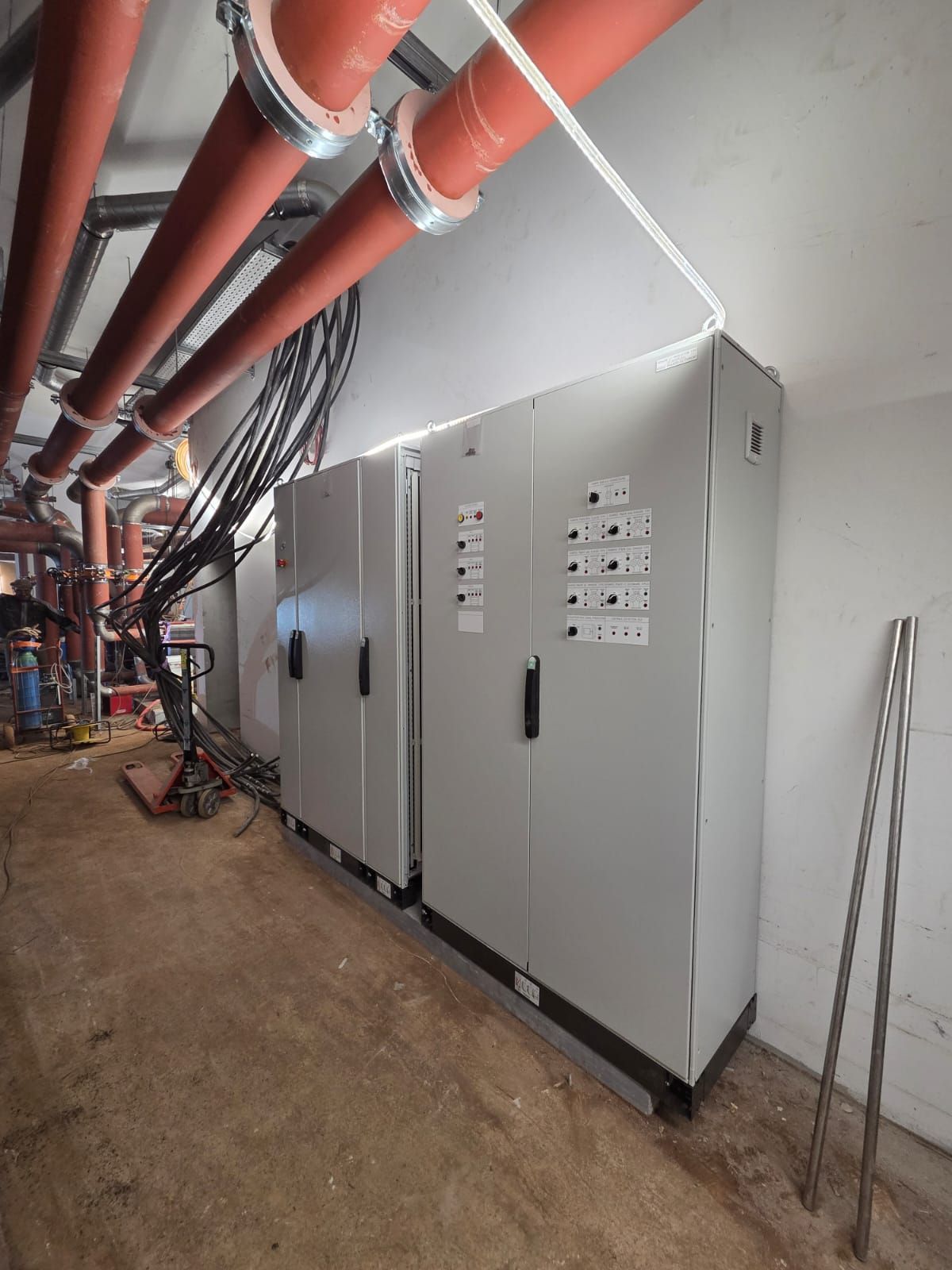 Grands armoires électriques de commande grises dans un couloir technique, avec des tuyaux rouges et des câbles au plafond