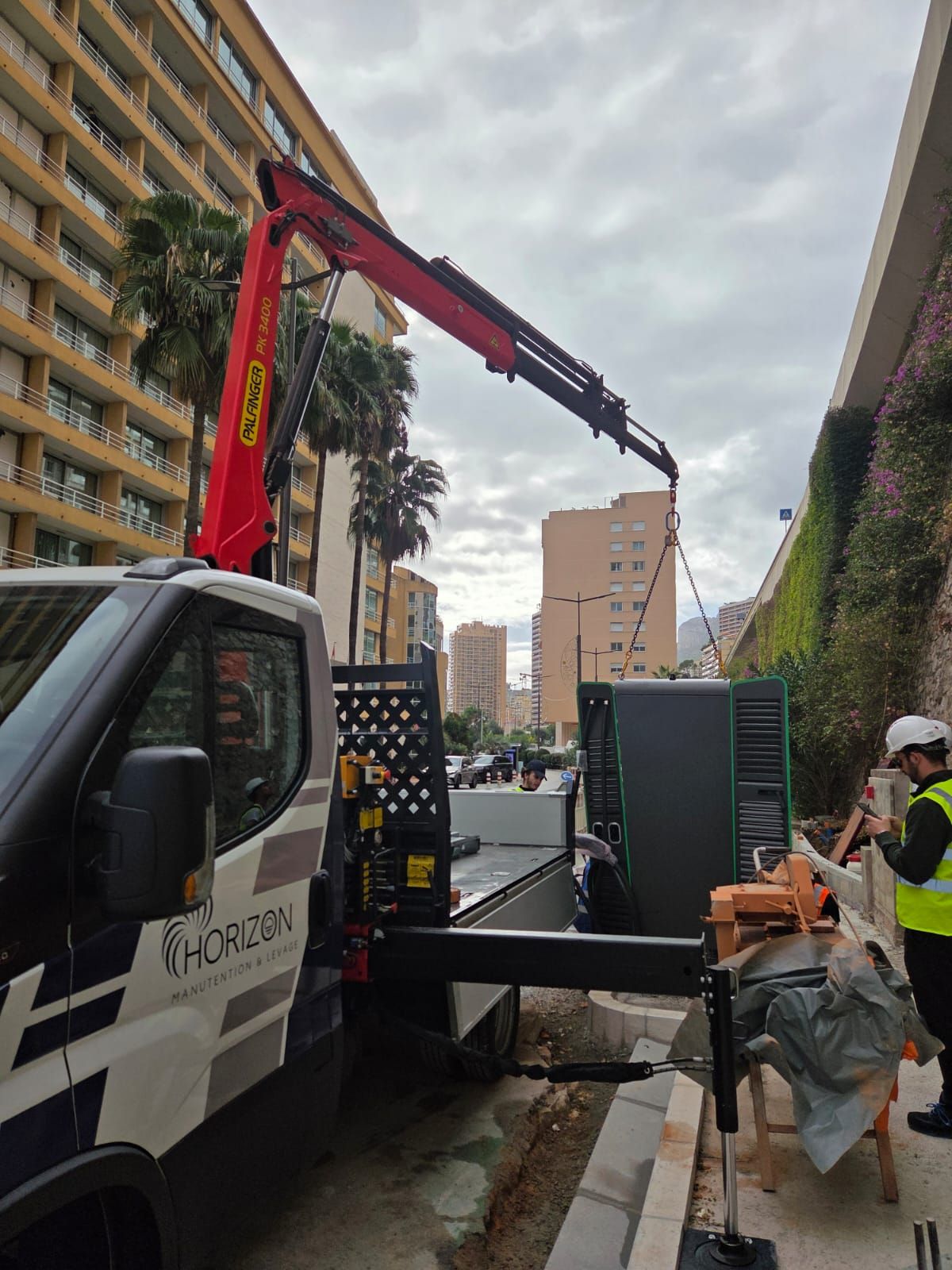Dépanneuse avec grue soulevant une grande benne noire dans une rue en ville, à côté d’immeubles résidentiels