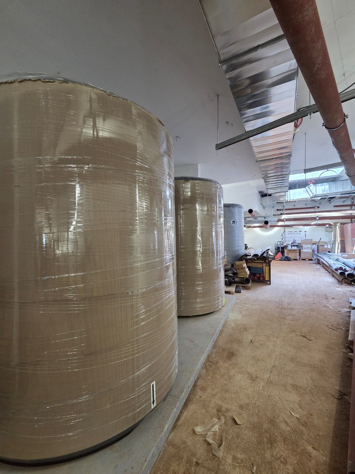 Des rangées de grands matériaux cylindriques emballés dans un couloir d’entrepôt en cours d’aménagement, avec des tuyaux apparents au plafond et des planches de bois au sol.