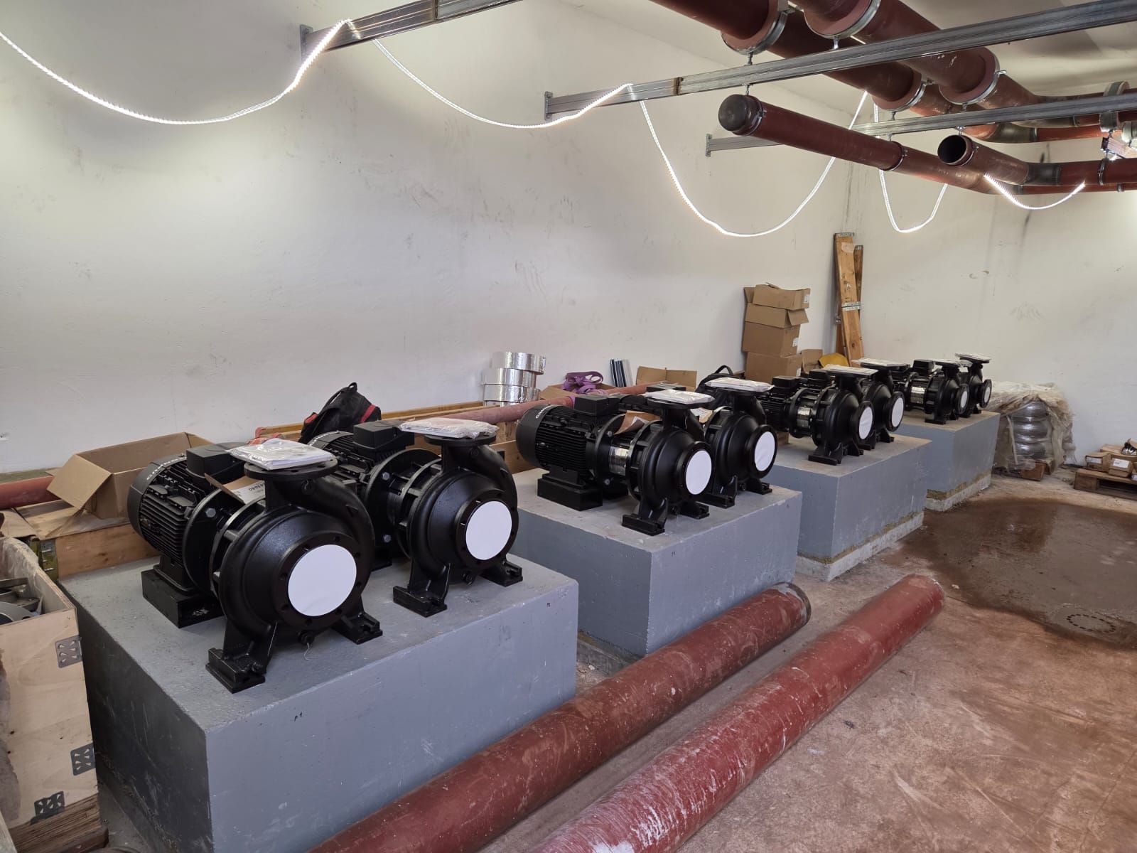 Rangée de pompes industrielles noires sur des socles en béton gris, dans une pièce de sous-sol en construction