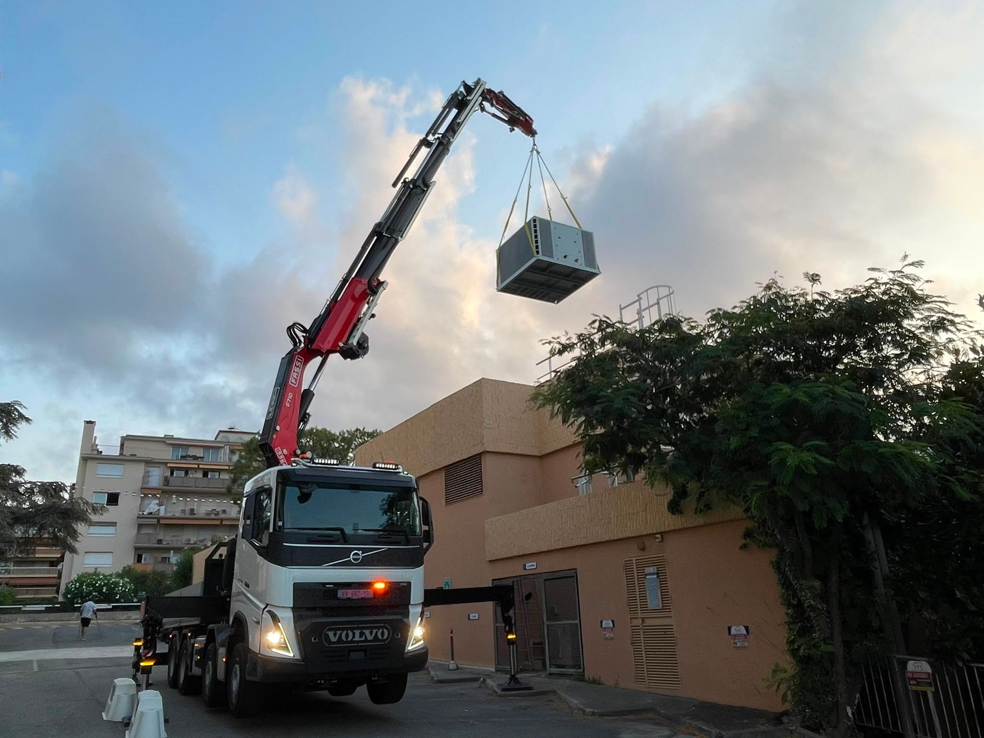 Un camion-grue soulève une petite structure au-dessus d’une rue bordée de bâtiments, au crépuscule