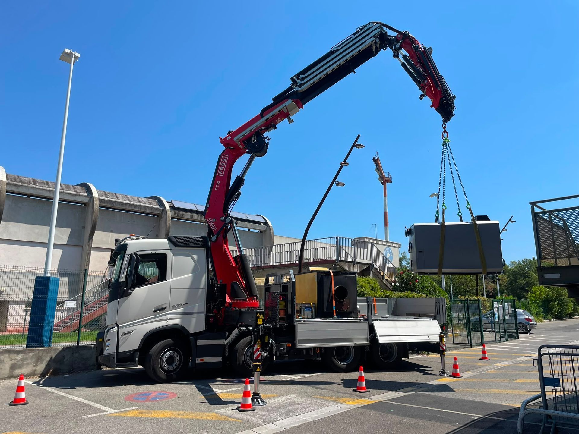 Un camion-grue soulève un grand conteneur noir dans une cour industrielle sous un ciel bleu