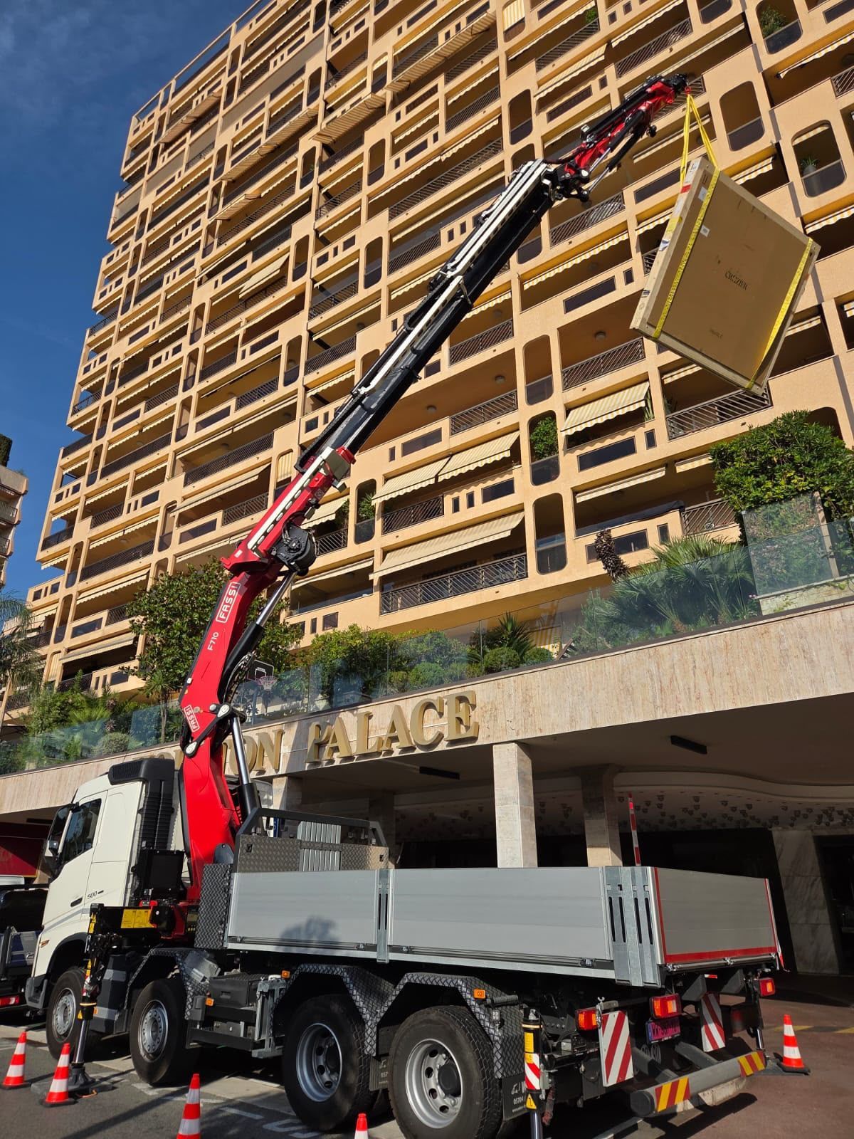 Une grue montée sur un camion soulève un grand panneau beige vers la façade d’un immeuble de grande hauteur couleur sable