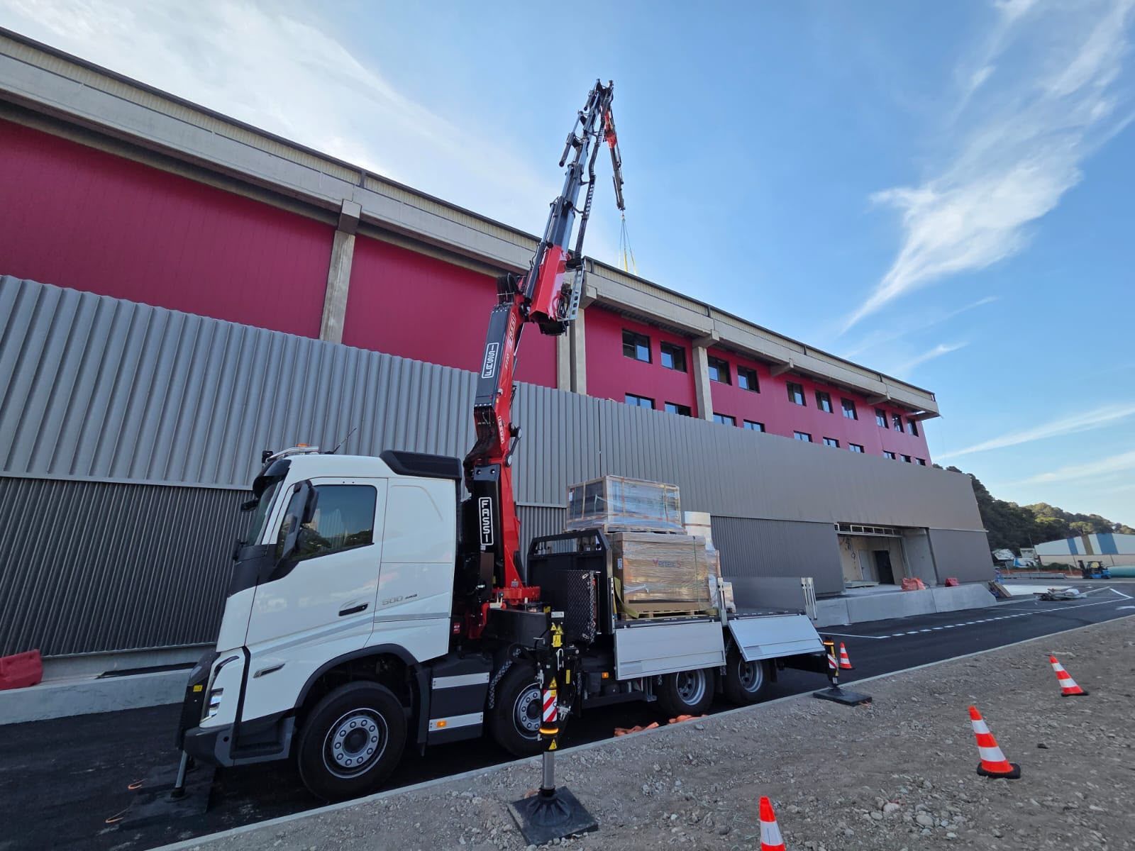 Un camion blanc équipé d’une grue charge des palettes à côté d’un entrepôt rose, sur le bas-côté en pente