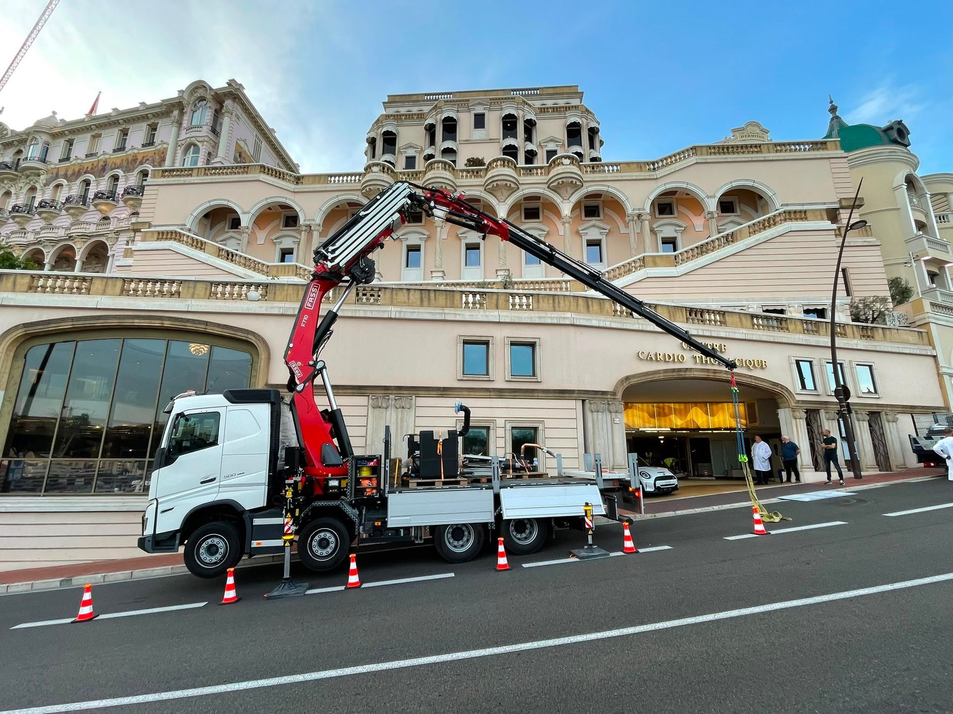 Camion-grue stationné devant un imposant bâtiment en pierre, sa flèche déployée au-dessus de cônes orange dans une rue de la ville.