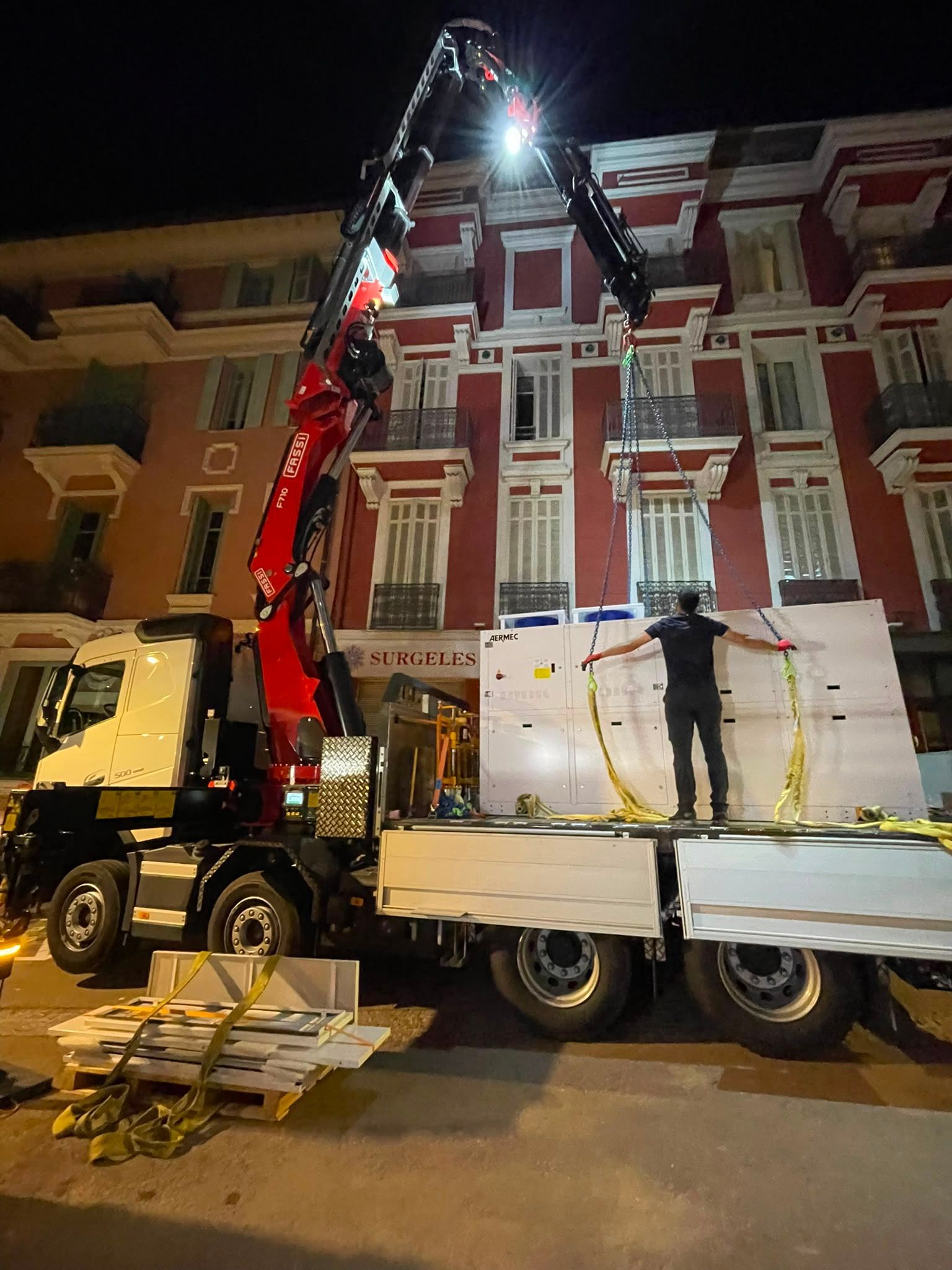 Une grue soulève une grosse charge blanche pour la déposer sur un camion, de nuit, devant des bâtiments colorés.