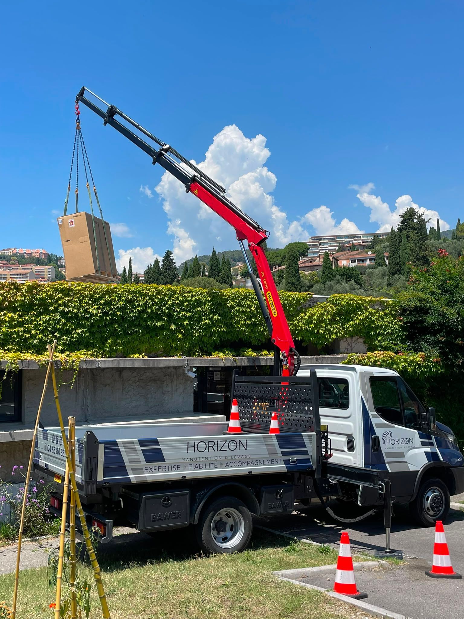 Un camion-grue soulève un gros objet au-dessus d’une haie, au bord d’une route, avec des cônes de signalisation et un autre camion à proximité.