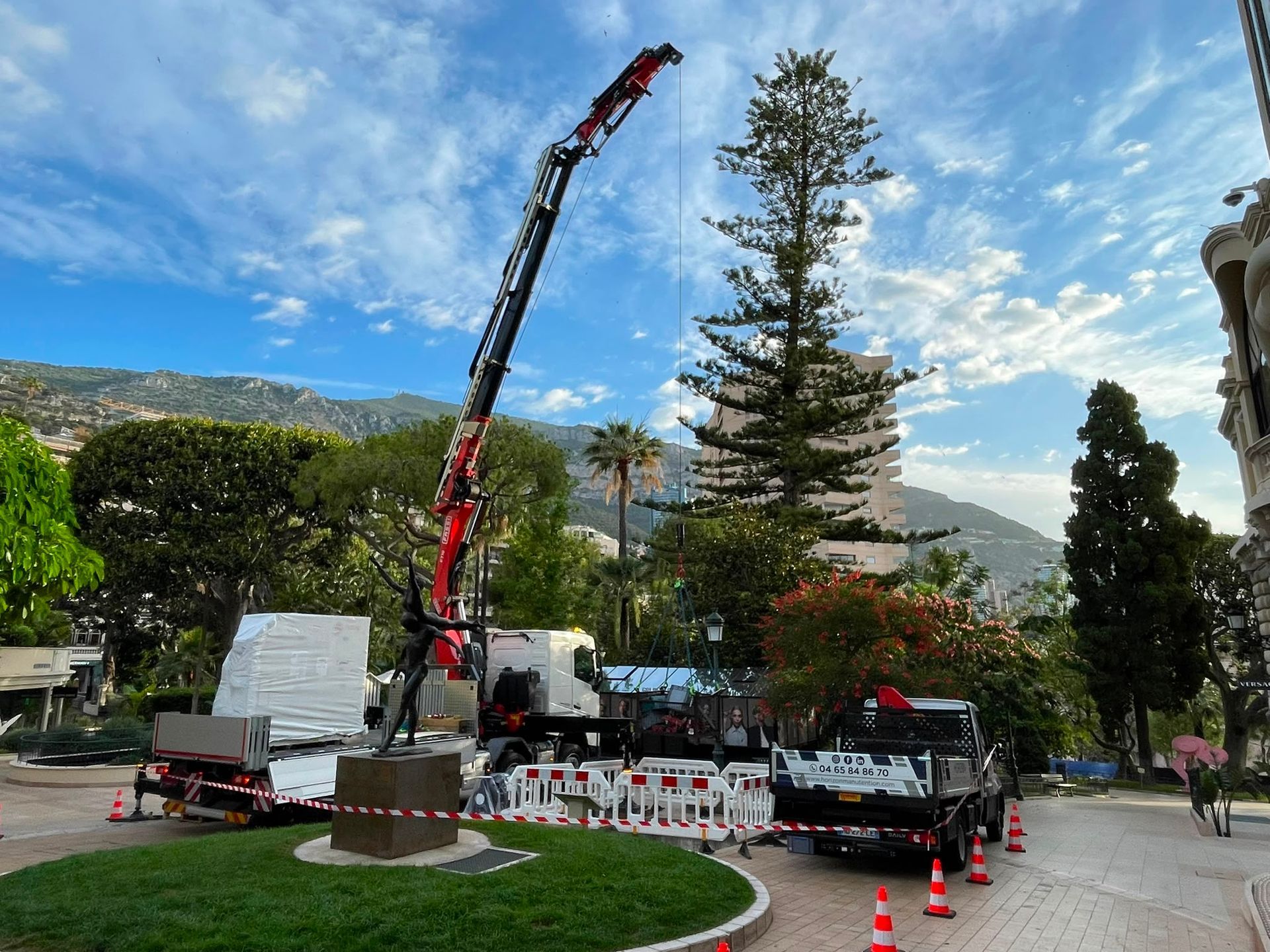 Une grue de chantier soulève des matériaux près de chaises blanches et de cônes, sur une place sous un ciel bleu