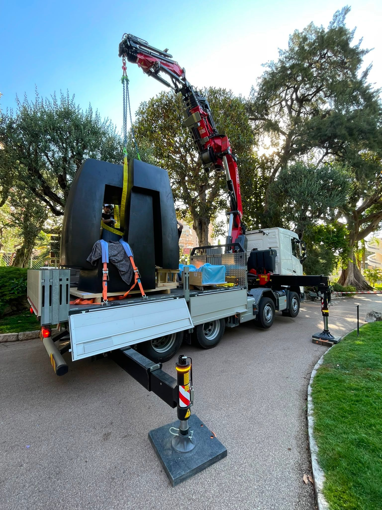 Grue montée sur camion soulevant une grande sculpture noire dans un cadre extérieur semblable à un parc