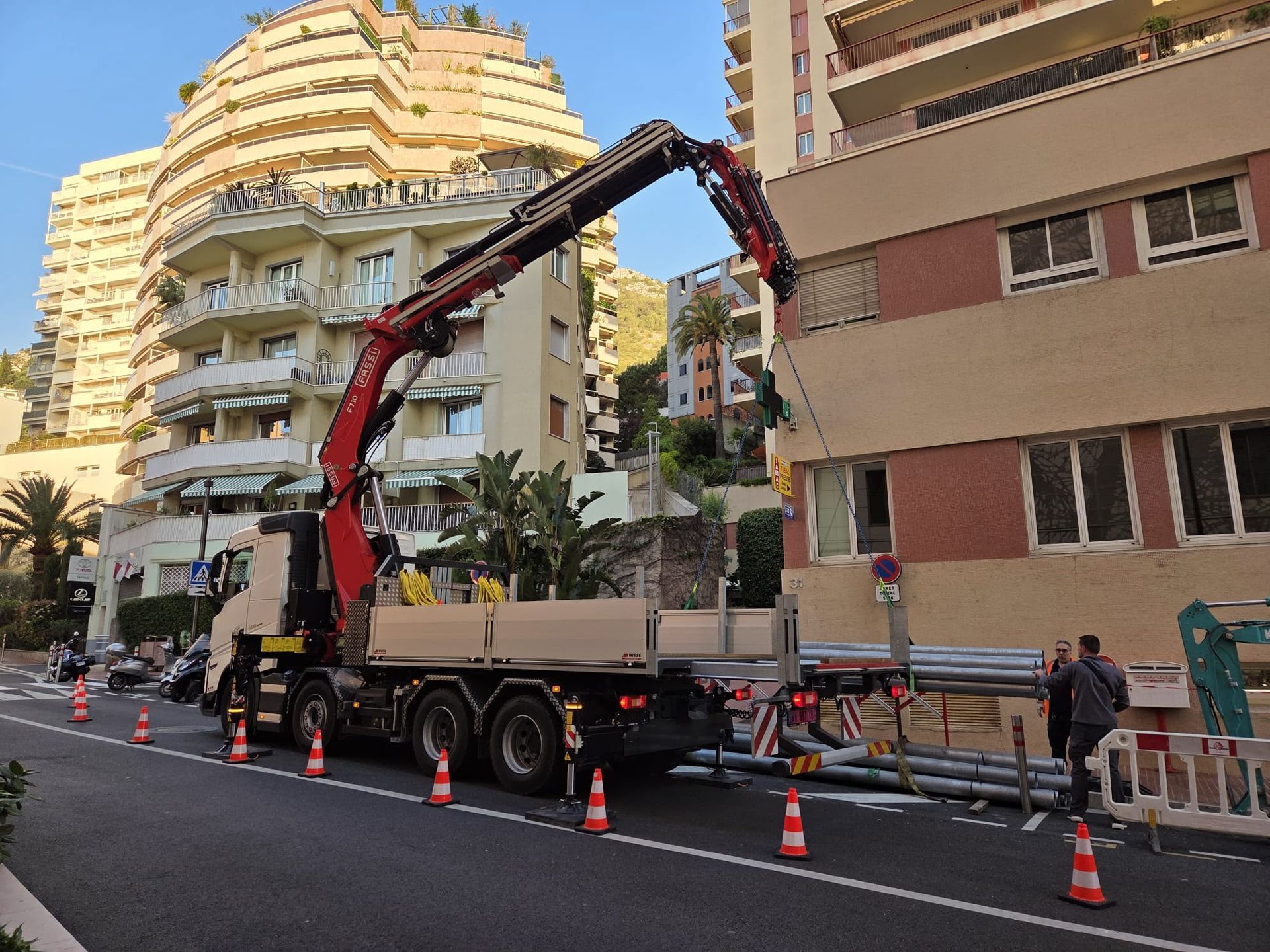Un camion-grue déchargeant des matériaux près d’un immeuble résidentiel en ville, avec des cônes de signalisation et des ouvriers à proximité