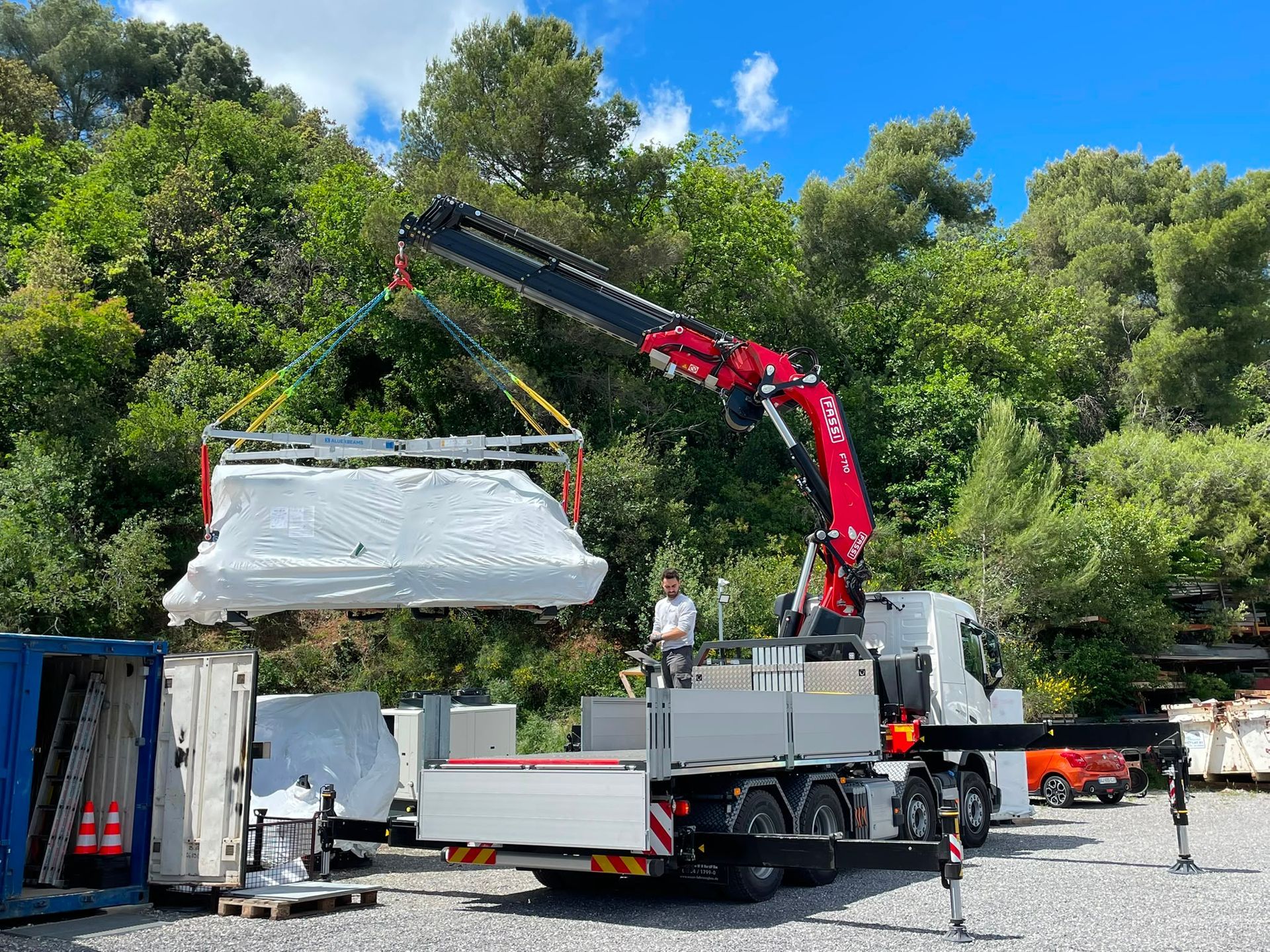 Une grue charge un gros objet blanc sur un camion, sur un chantier en plein air dans une zone boisée