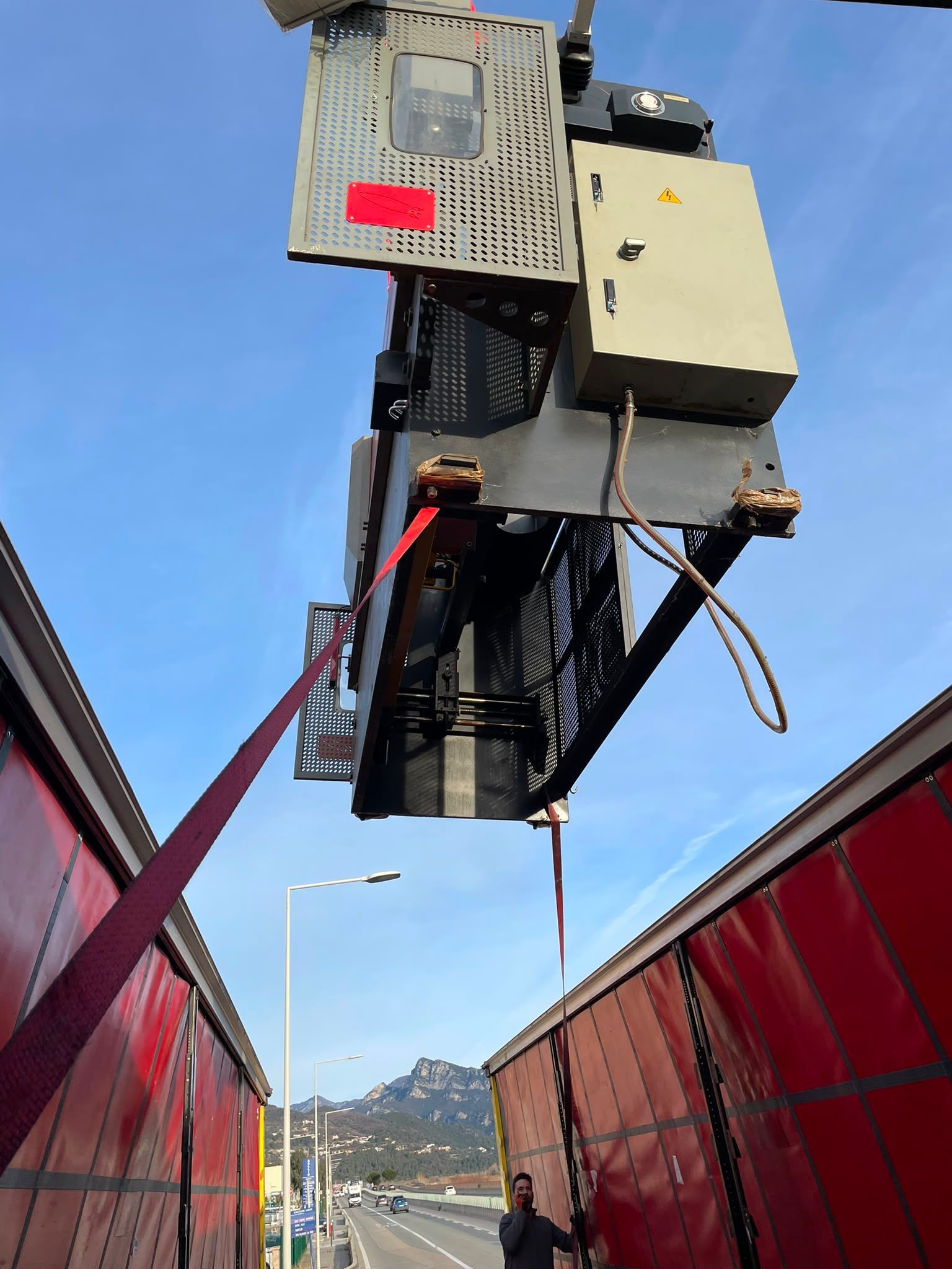 Machine de maintenance suspendue sur un rail entre des panneaux rouges, sur fond de ciel bleu, avec une route en contrebas.