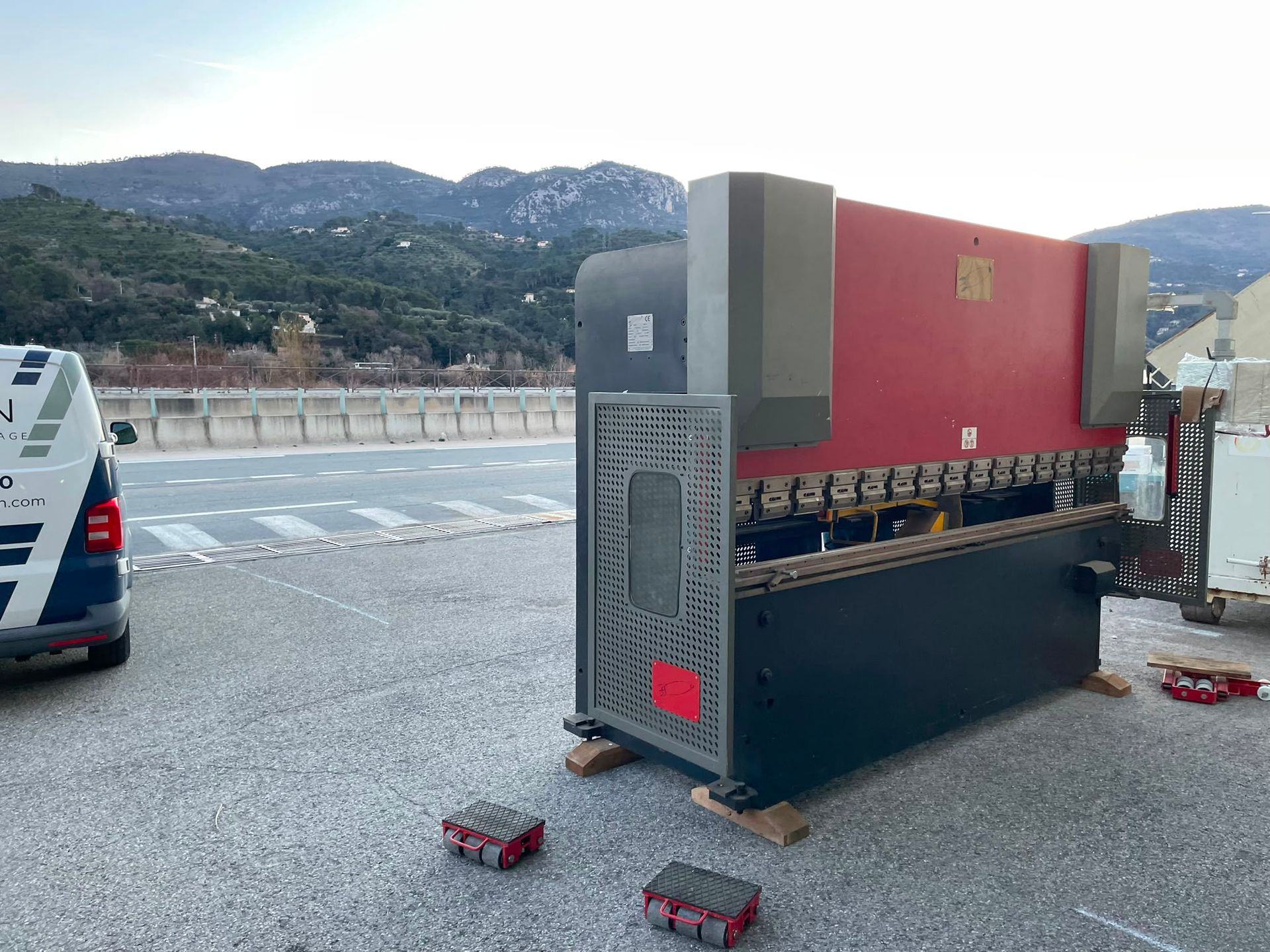 Machine rouge renversée sur du gravier au bord d’une route, avec une camionnette et des montagnes en arrière-plan