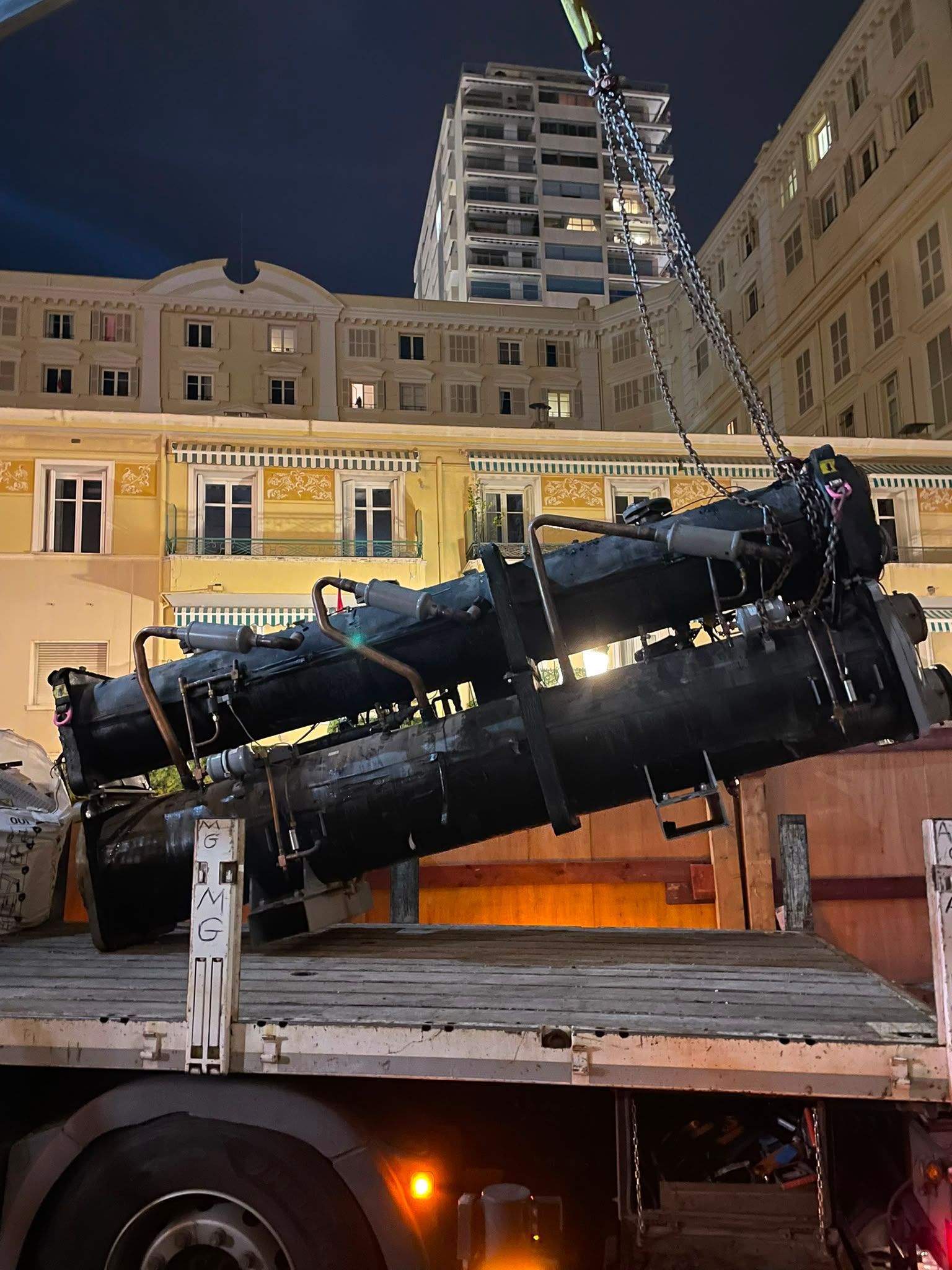 Grand télescope noir installé à l’arrière d’un camion au crépuscule, avec des immeubles et une grue en arrière-plan