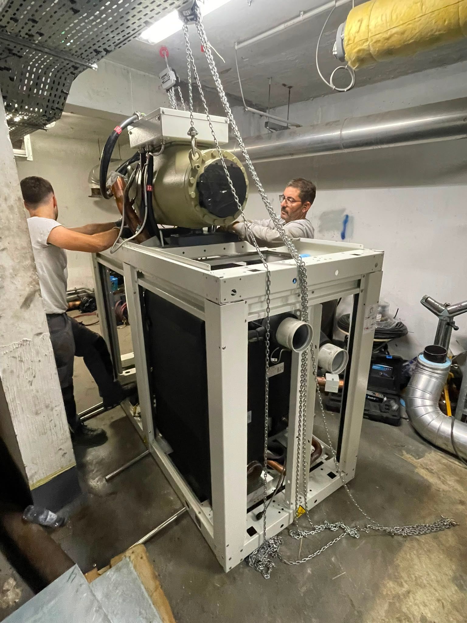 Deux ouvriers manœuvrent un gros équipement de chauffage, ventilation et climatisation ou un appareil industriel dans un local technique où les conduits et les tuyaux sont apparents