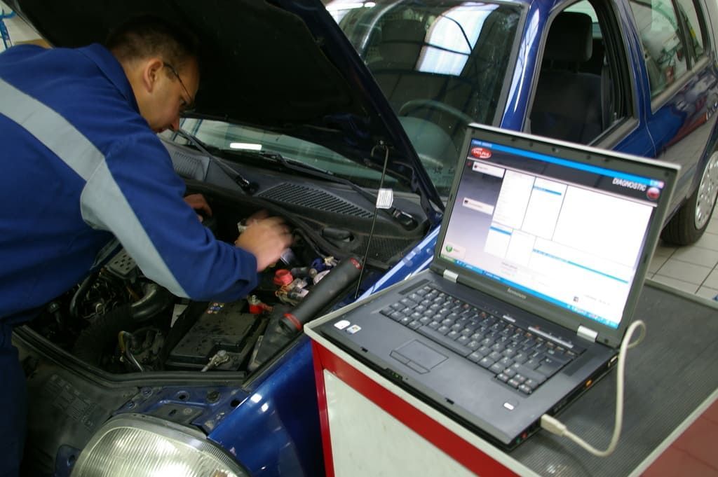 Un mécanicien travaille sous le capot d'une voiture, à côté d'un ordinateur portable affichant des diagnostics.