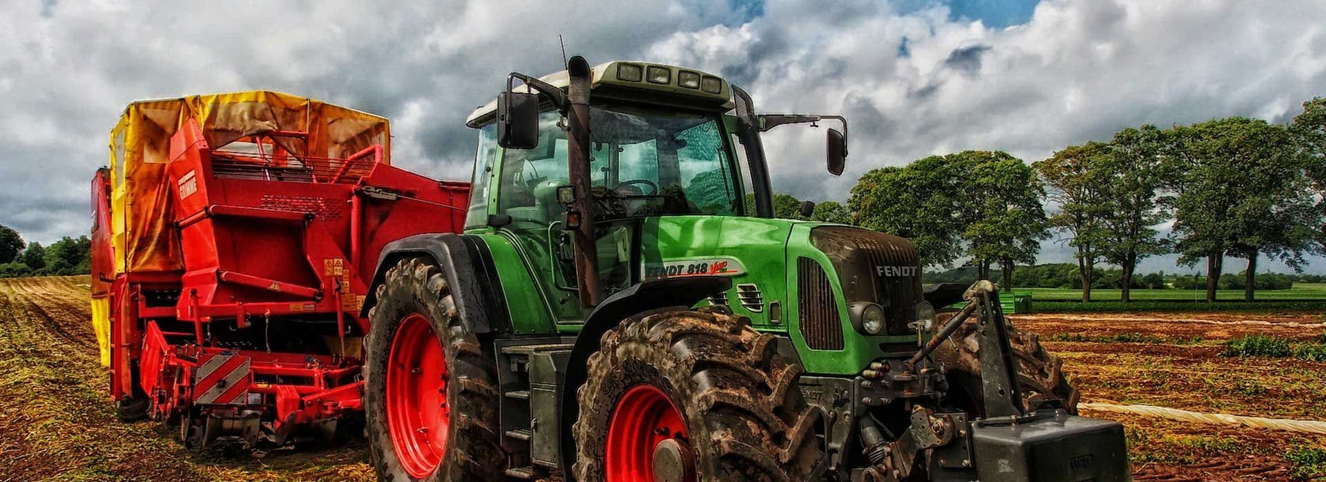 Un tracteur vert tirant un outil agricole rouge dans un champ boueux sous un ciel nuageux.