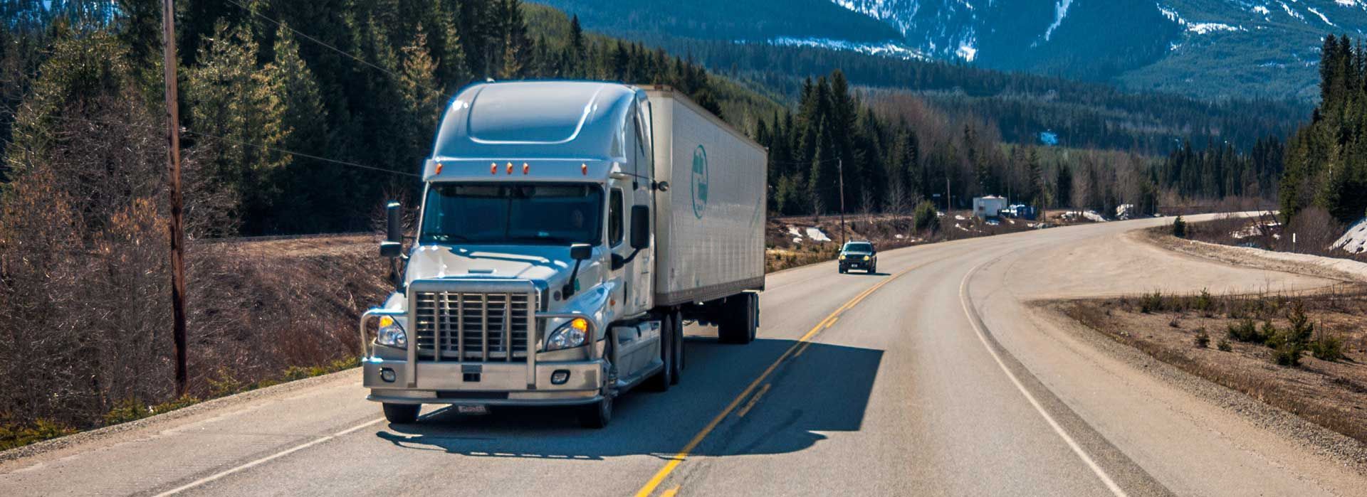 Un semi-remorque bleu circule sur une route de montagne avec des sommets enneigés et des pins en arrière-plan.