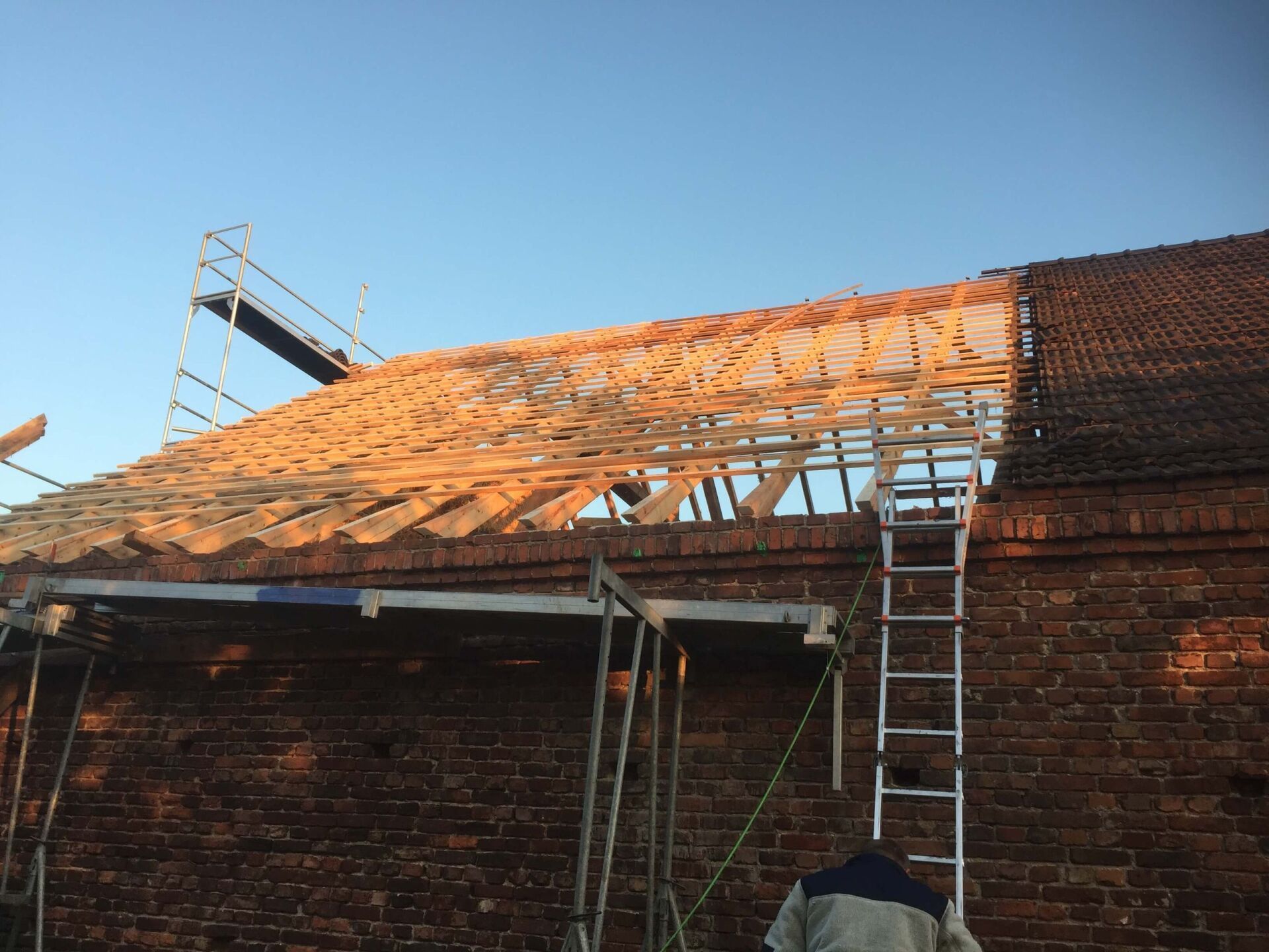 Teilweise repariertes Dach mit freiliegenden Holzsparren und einer an ein Backsteingebäude gelehnten Leiter.