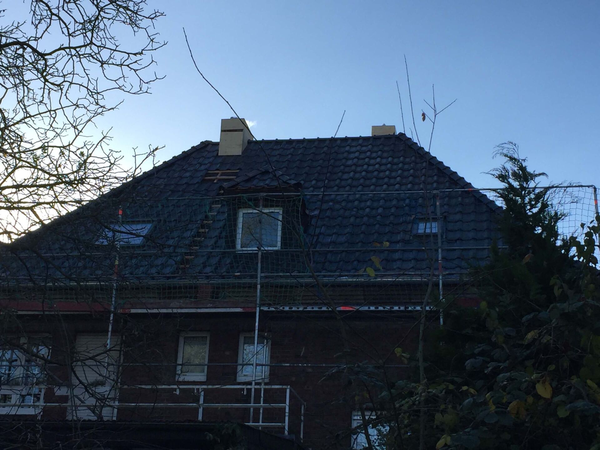 Dunkles Haus mit steilem Ziegeldach, von unten zwischen kahlen Bäumen und Stromleitungen gesehen.