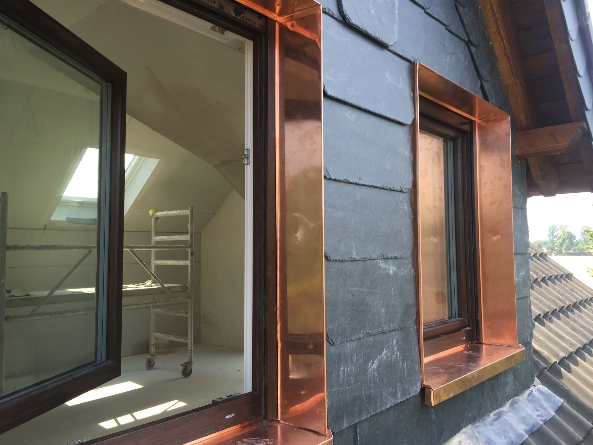 Kupferverzierte Dachgauben an einem schiefergedeckten Haus, wobei ein Fenster zu einem Innenraum hin geöffnet ist.
