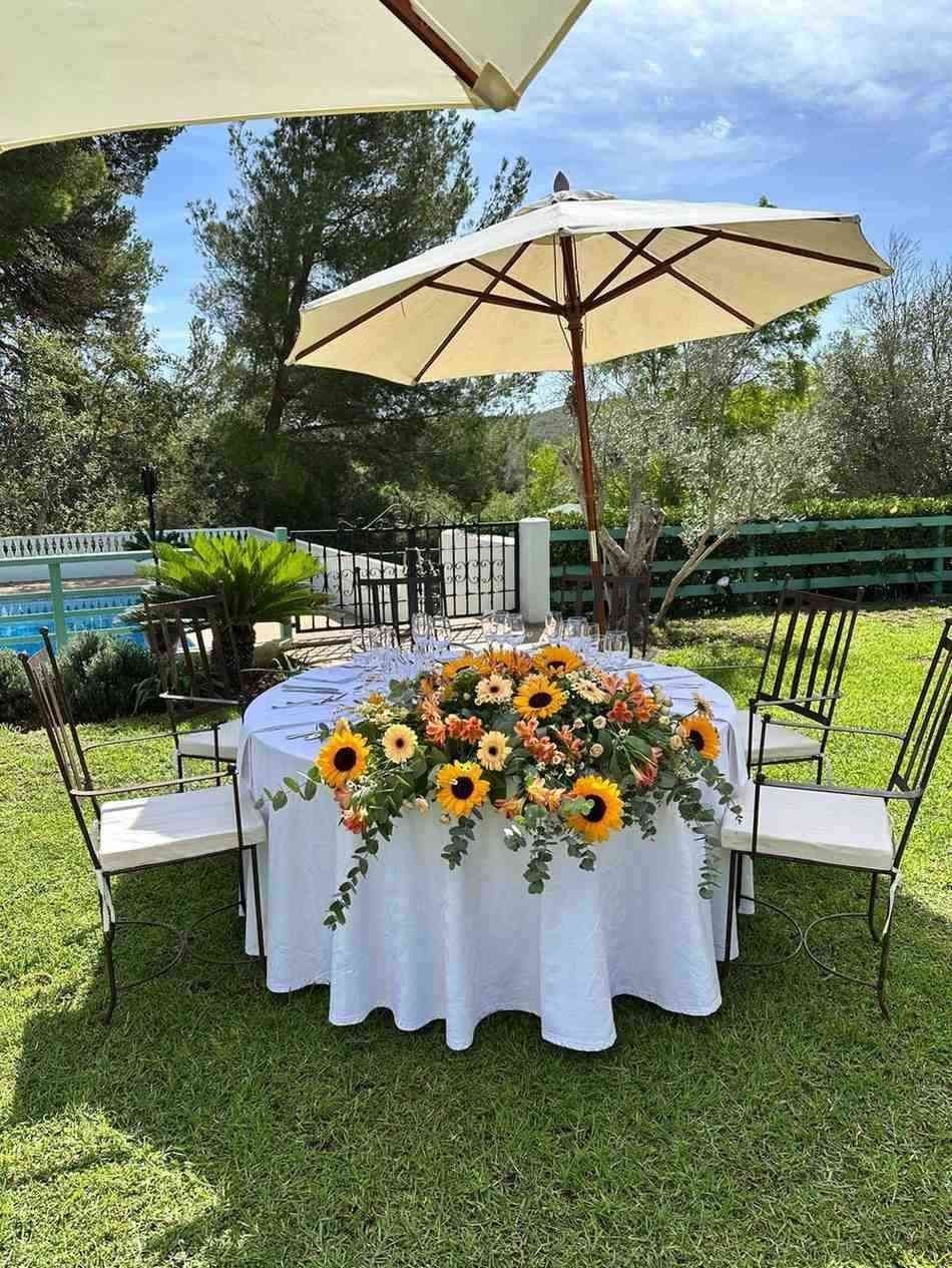 Una mesa redonda con mantel blanco y girasoles, colocada al aire libre bajo una sombrilla beige en un césped soleado.
