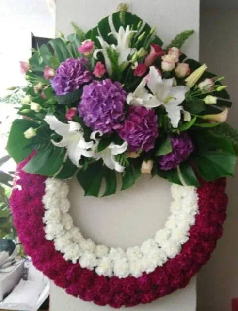 Una corona fúnebre con un aro de claveles rojos y blancos, rematada con un vibrante arreglo de hortensias y lirios morados.