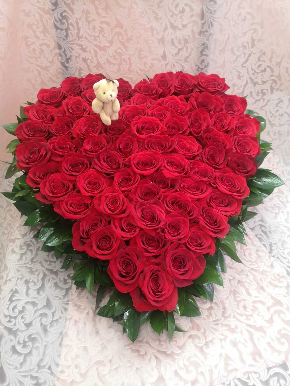 Un gran arreglo floral en forma de corazón, compuesto por vibrantes rosas rojas y adornado con un pequeño osito de peluche, sobre un fondo floral blanco.