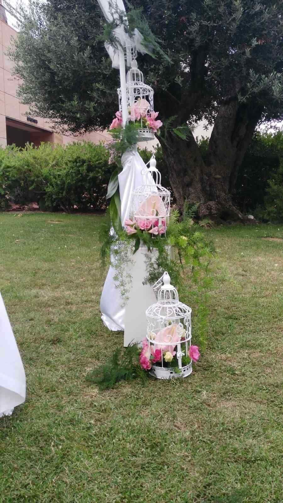 En un jardín cubierto de césped, se exhiben jaulas decorativas blancas para pájaros, llenas de flores rosas y vegetación, sobre un soporte.