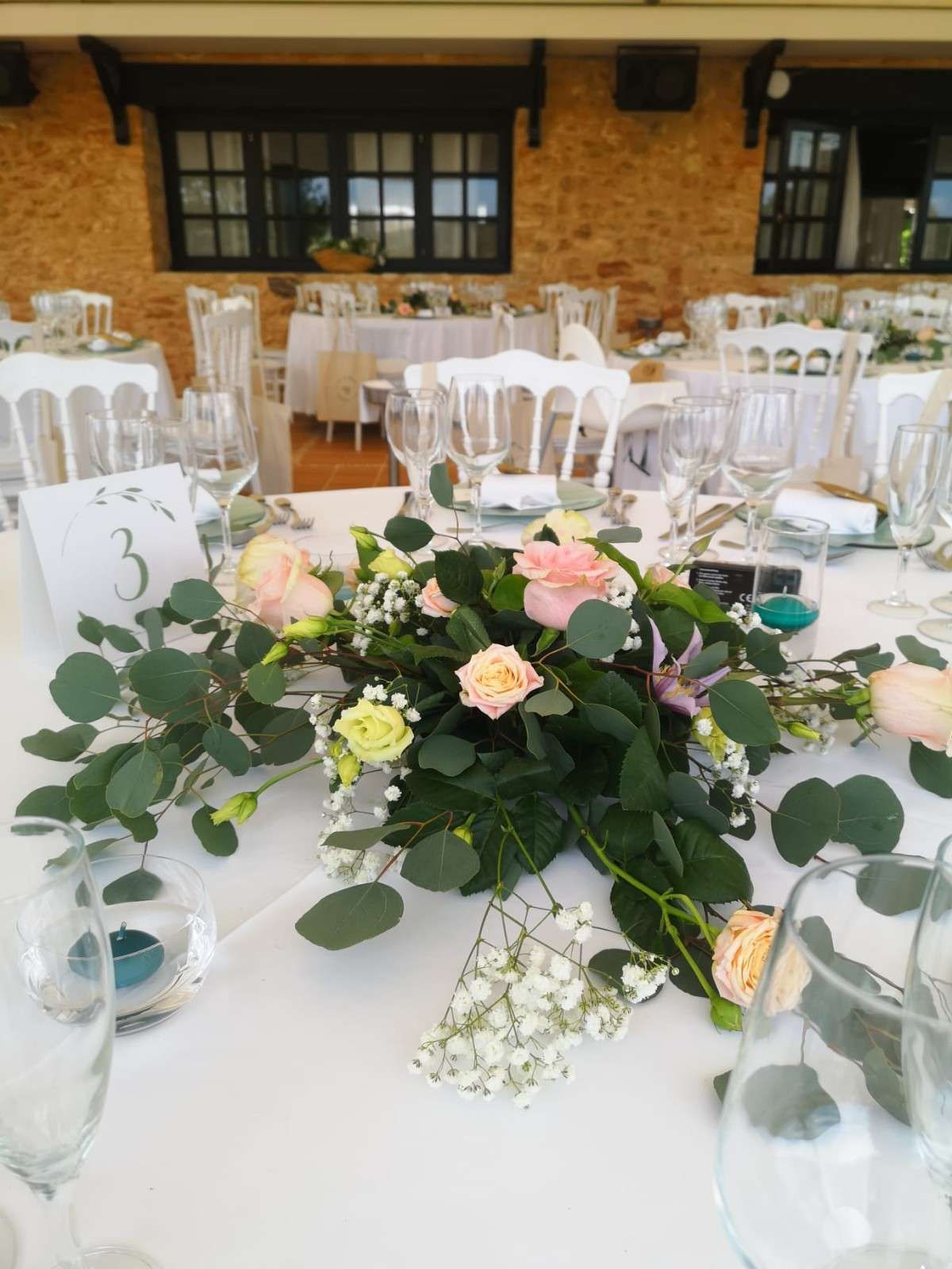 Un centro de mesa floral con rosas rosadas y amarillas y eucalipto sobre una mesa blanca preparada para un evento, con el número 3 visible.