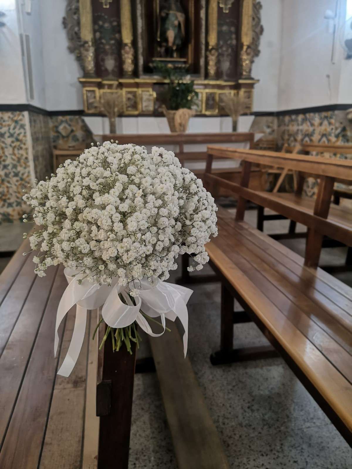 Un ramo de flores blancas de paniculata atado con una cinta blanca a un banco de madera de una iglesia, con un altar al fondo.
