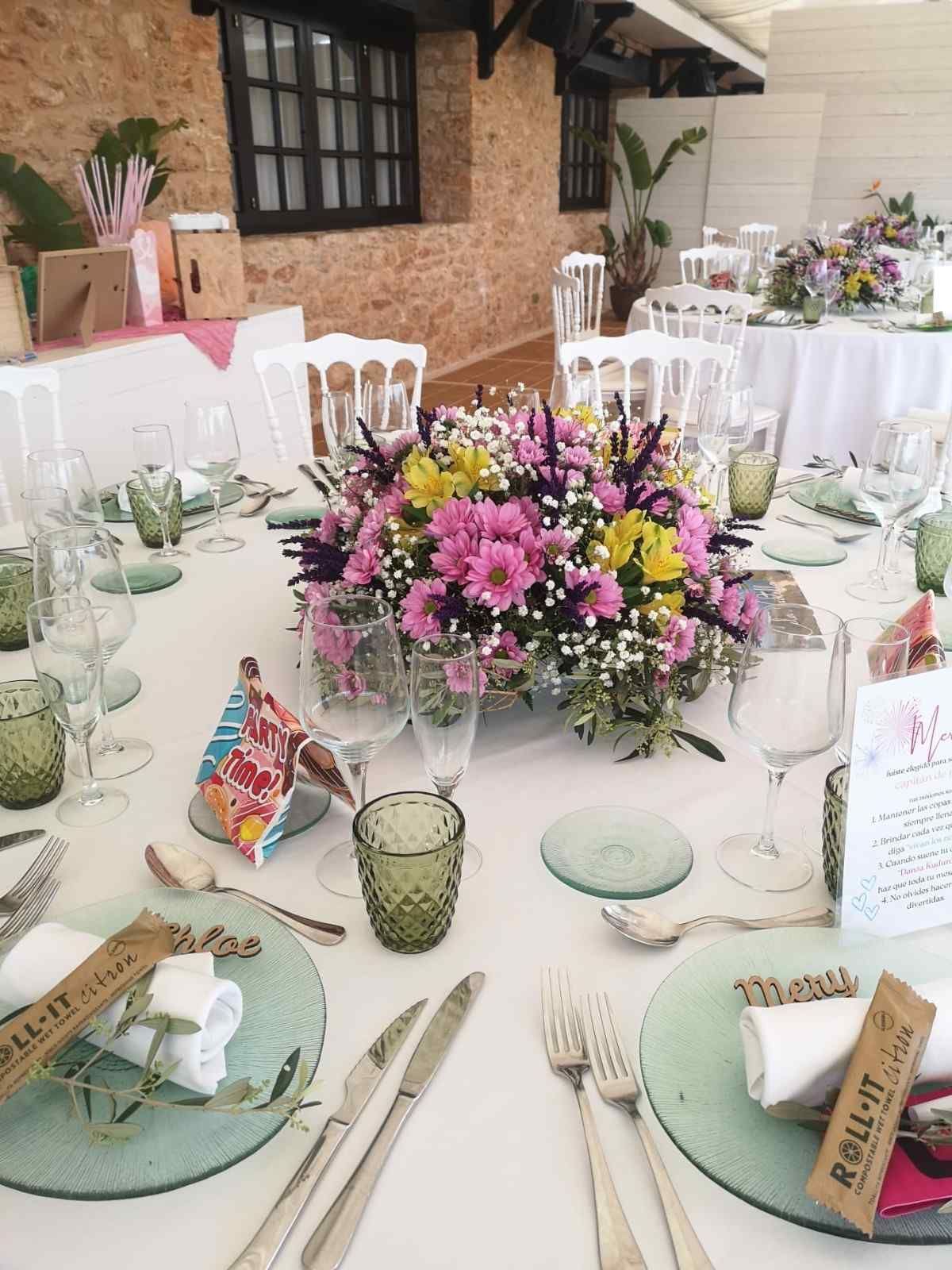 Una mesa nupcial puesta con un colorido centro de mesa floral, mantelería blanca, cristalería verde y cubiertos con menús.
