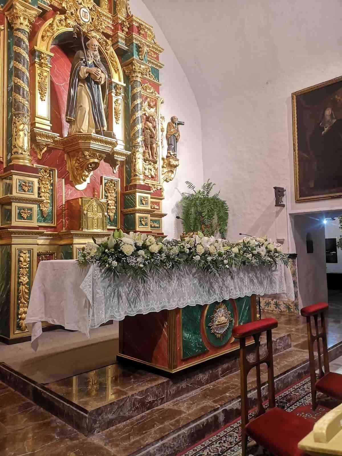 Un altar dorado ornamentado en una iglesia decorada con flores blancas, con dos bancos de madera para arrodillarse en primer plano.