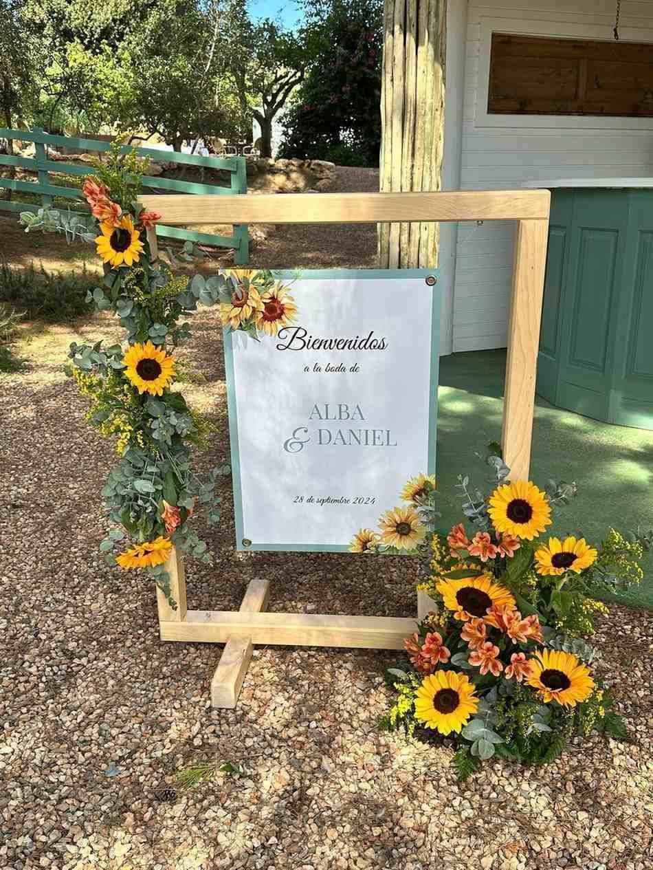 Un cartel de bienvenida de madera para la boda de Alma y Daniel, adornado con girasoles y eucalipto, situado sobre un camino de grava.