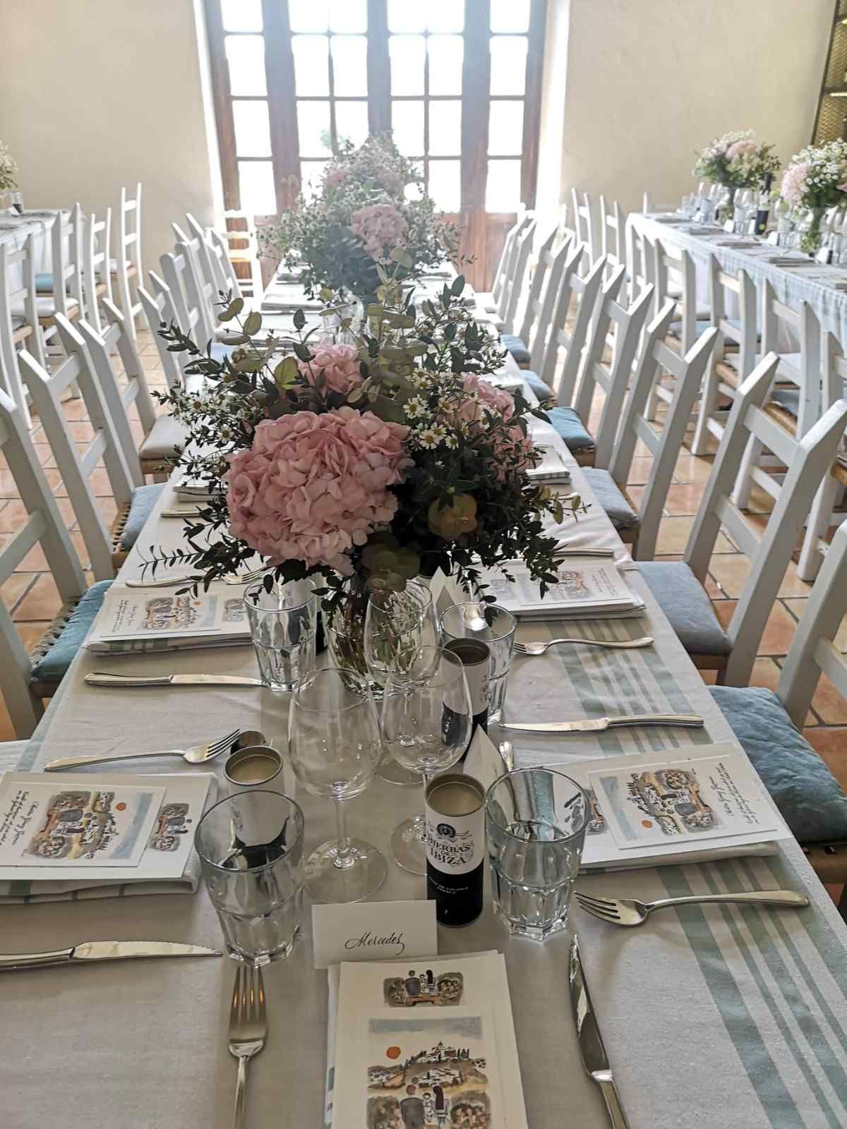 Una larga mesa de banquete preparada para una boda con centros de mesa florales rosas, tarjetas de sitio blancas y menús en una sala luminosa.