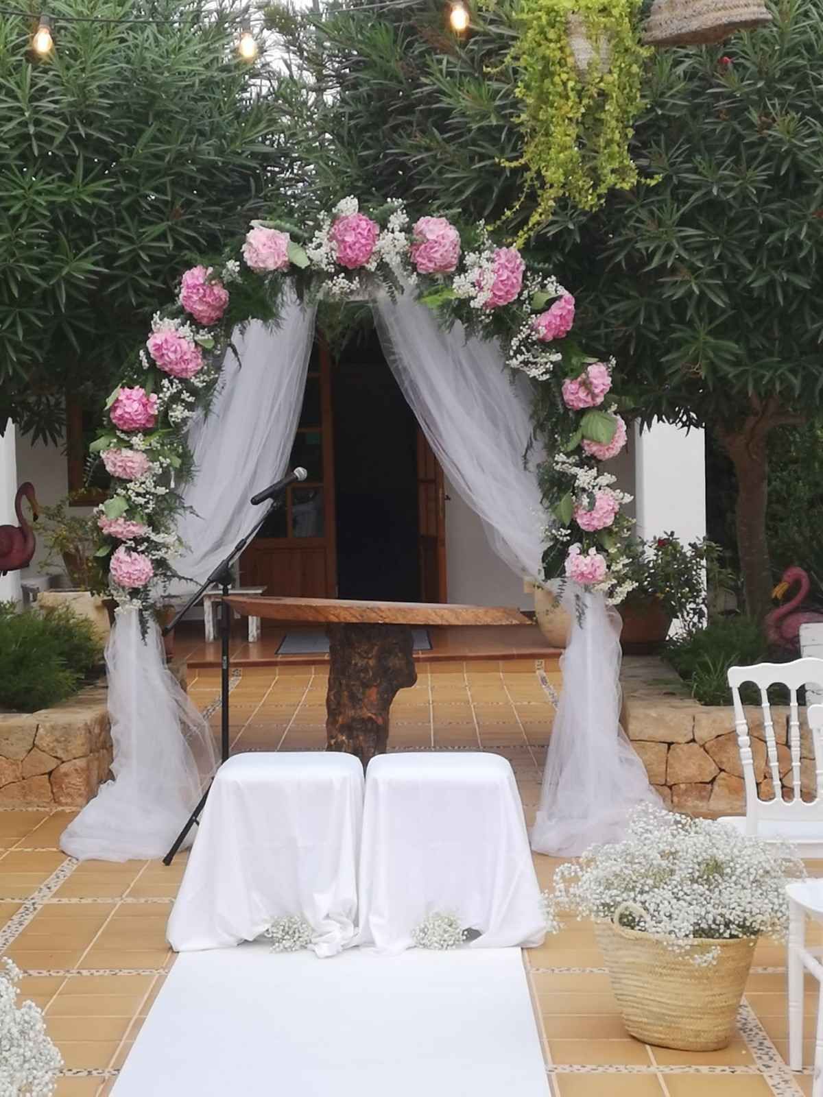 Un arco nupcial adornado con flores rosas y tela blanca se alza sobre una mesa rústica de madera y dos taburetes blancos.