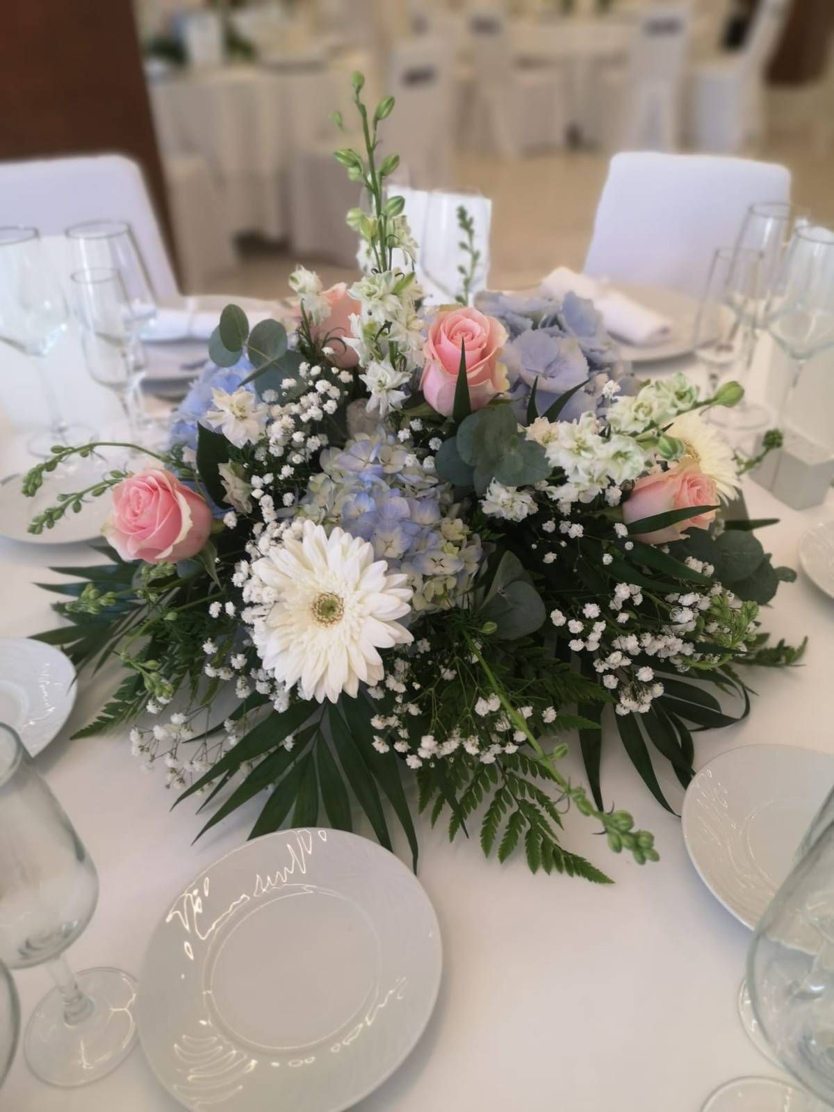 Un centro de mesa floral con rosas rosadas, una gerbera blanca, hortensias azules y follaje sobre un mantel blanco.