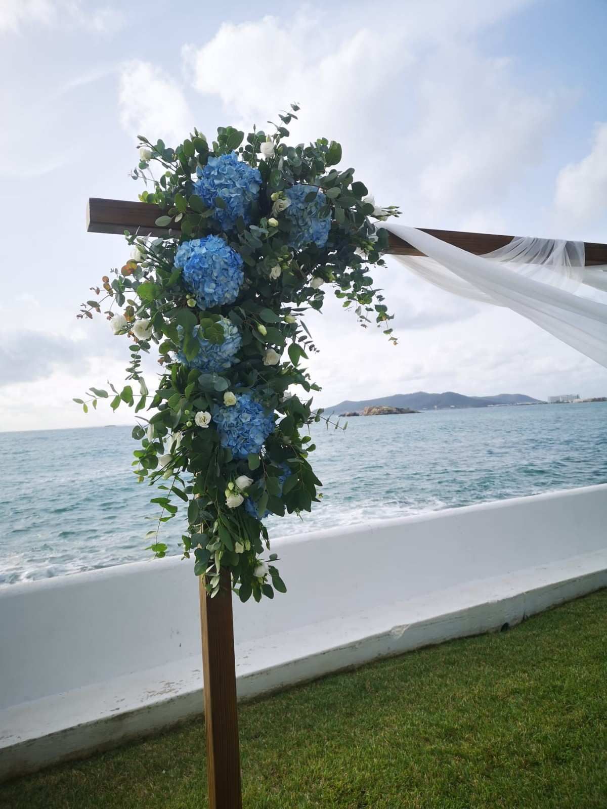 Arreglo floral de hortensias azules y follaje verde sobre un arco de madera para bodas con vistas al océano.