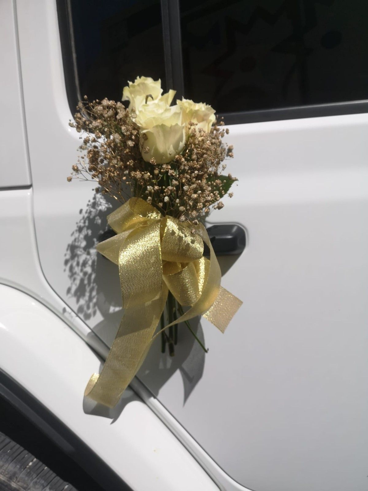 Un ramo decorativo de rosas blancas y paniculata, atado con una cinta dorada, sujeto a la manija blanca de la puerta de un coche.