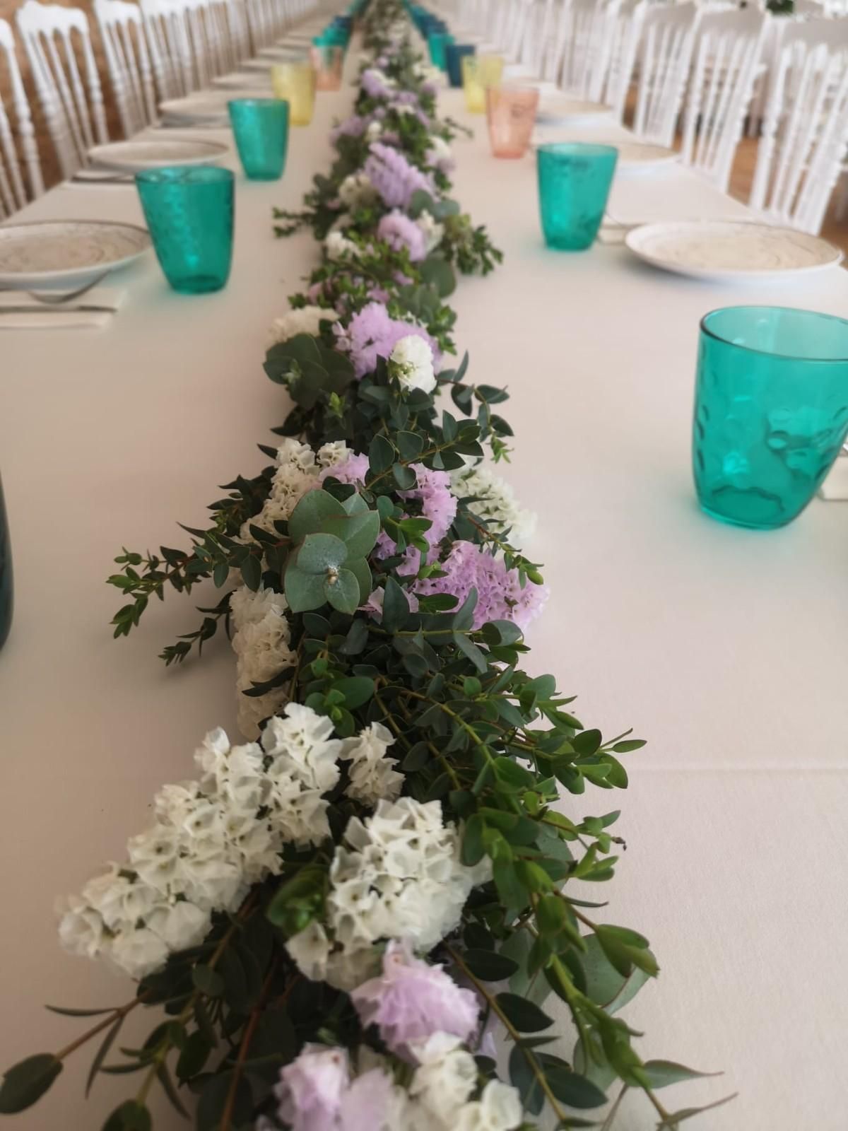 Una larga mesa de banquete puesta con manteles blancos, sillas, platos y vasos de color verde azulado, decorada con un camino de mesa de follaje floral.