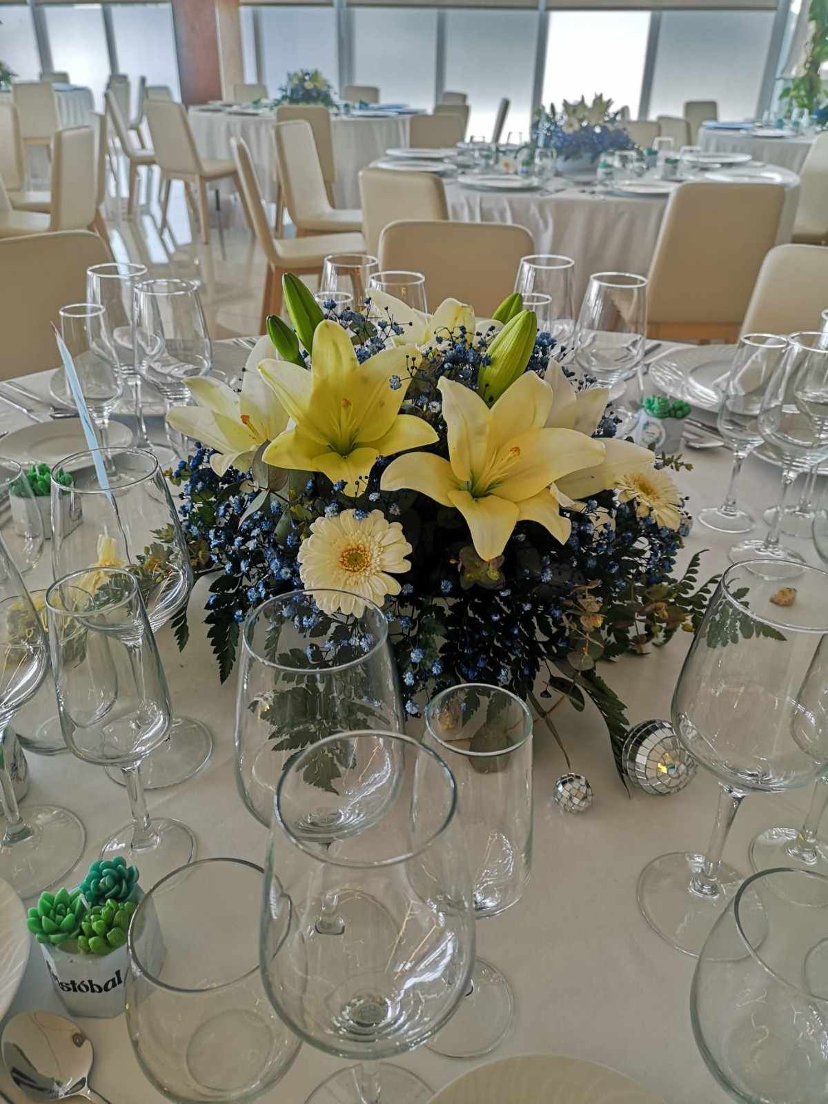 Un centro de mesa floral con lirios color crema y flores azul oscuro sobre una mesa cubierta con un mantel blanco, rodeada de copas de vino vacías.