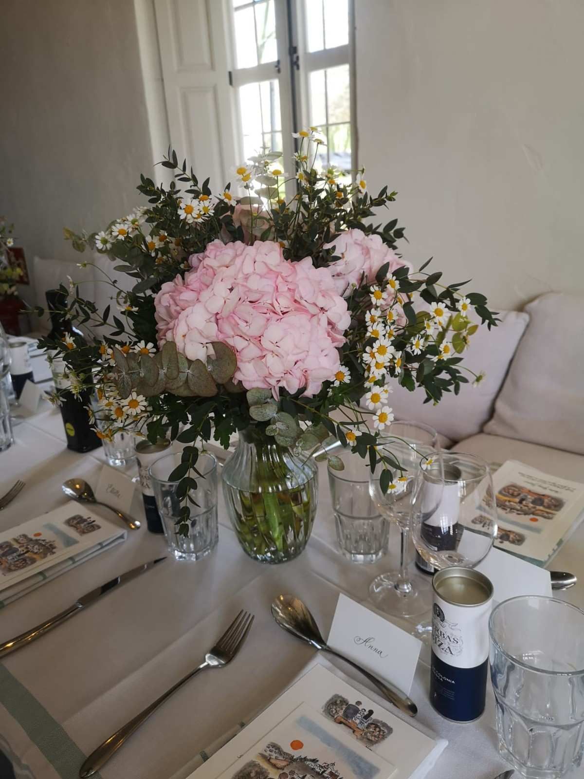 Un centro de mesa con hortensias rosas y margaritas blancas en un jarrón transparente, rodeado de vajilla.