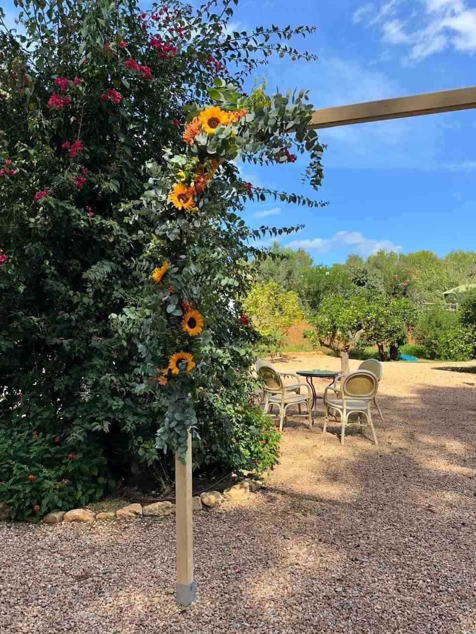 Un arco de madera adornado con hojas de eucalipto y girasoles se alza en un jardín al aire libre con asientos en el patio.