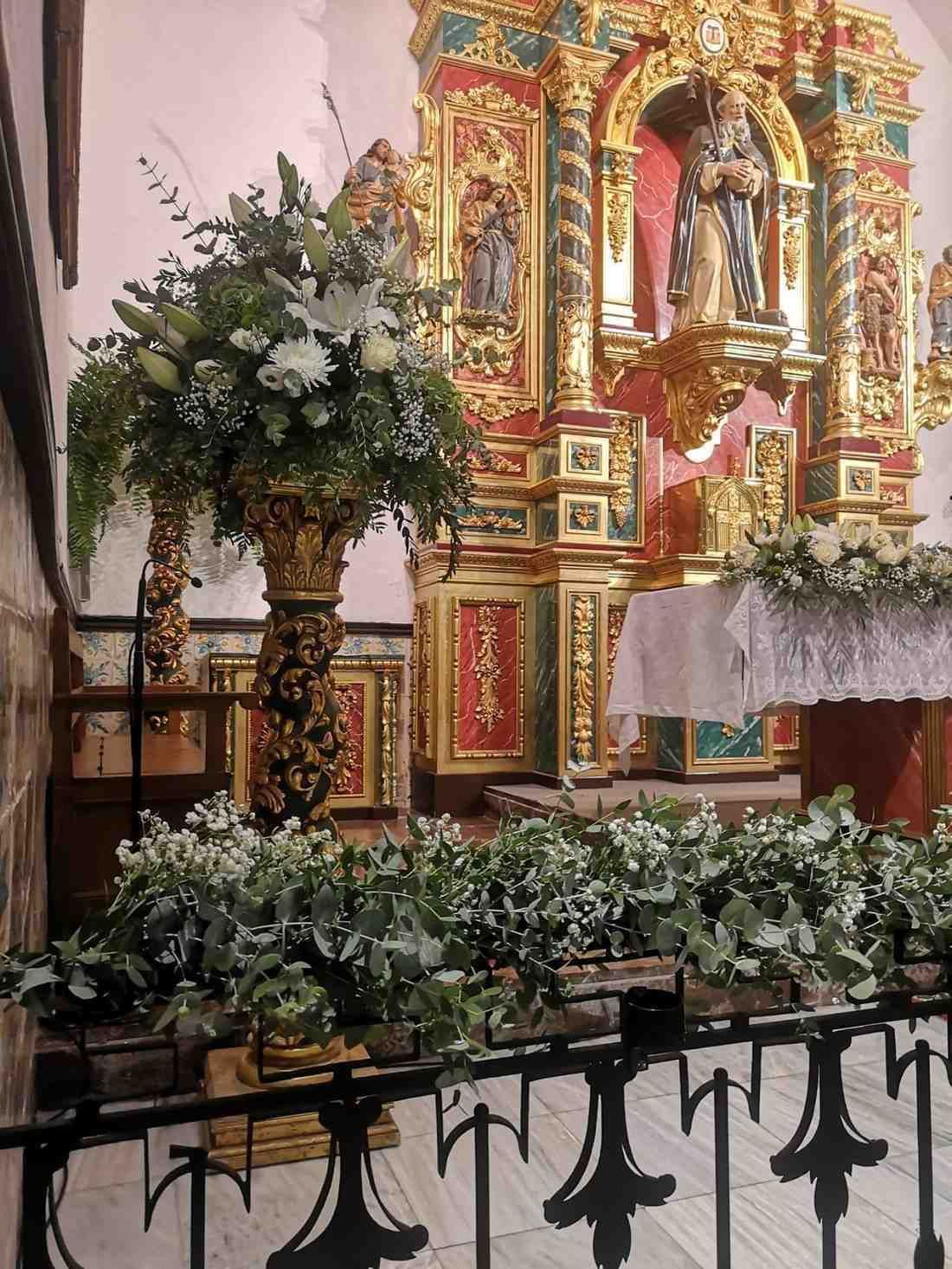 Un arreglo floral reposa sobre un alto y ornamentado pedestal dorado dentro de una iglesia, con un altar dorado al fondo.